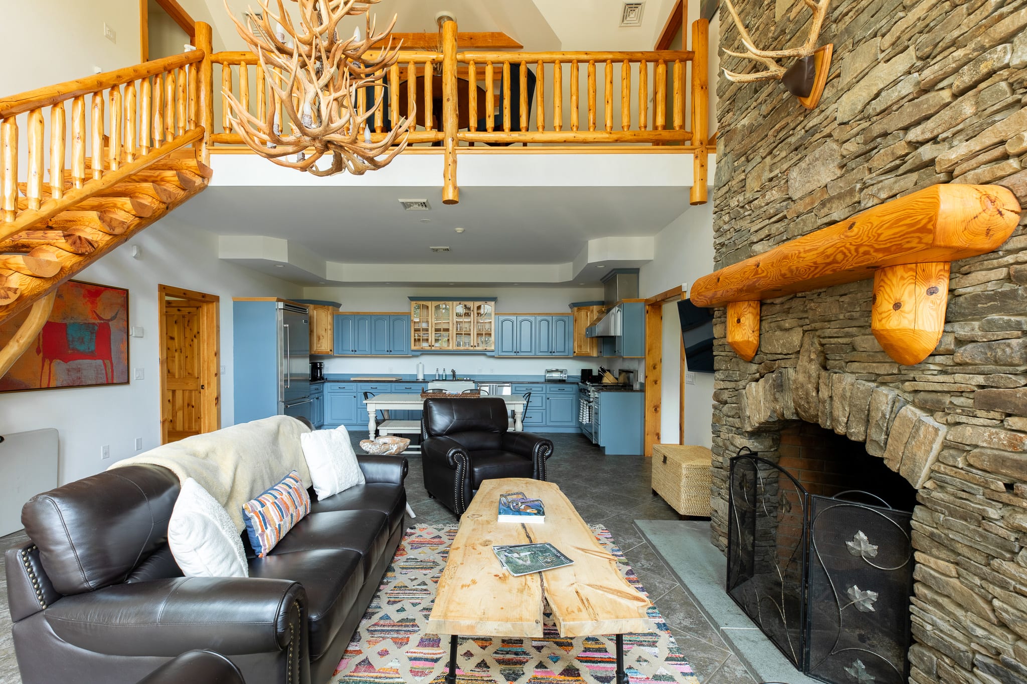 An open-concept living space featuring a large stone fireplace, a leather sofa, and a rustic wood coffee table, with a blue kitchen visible in the background and a loft area above. An antler chandelier hangs prominently.