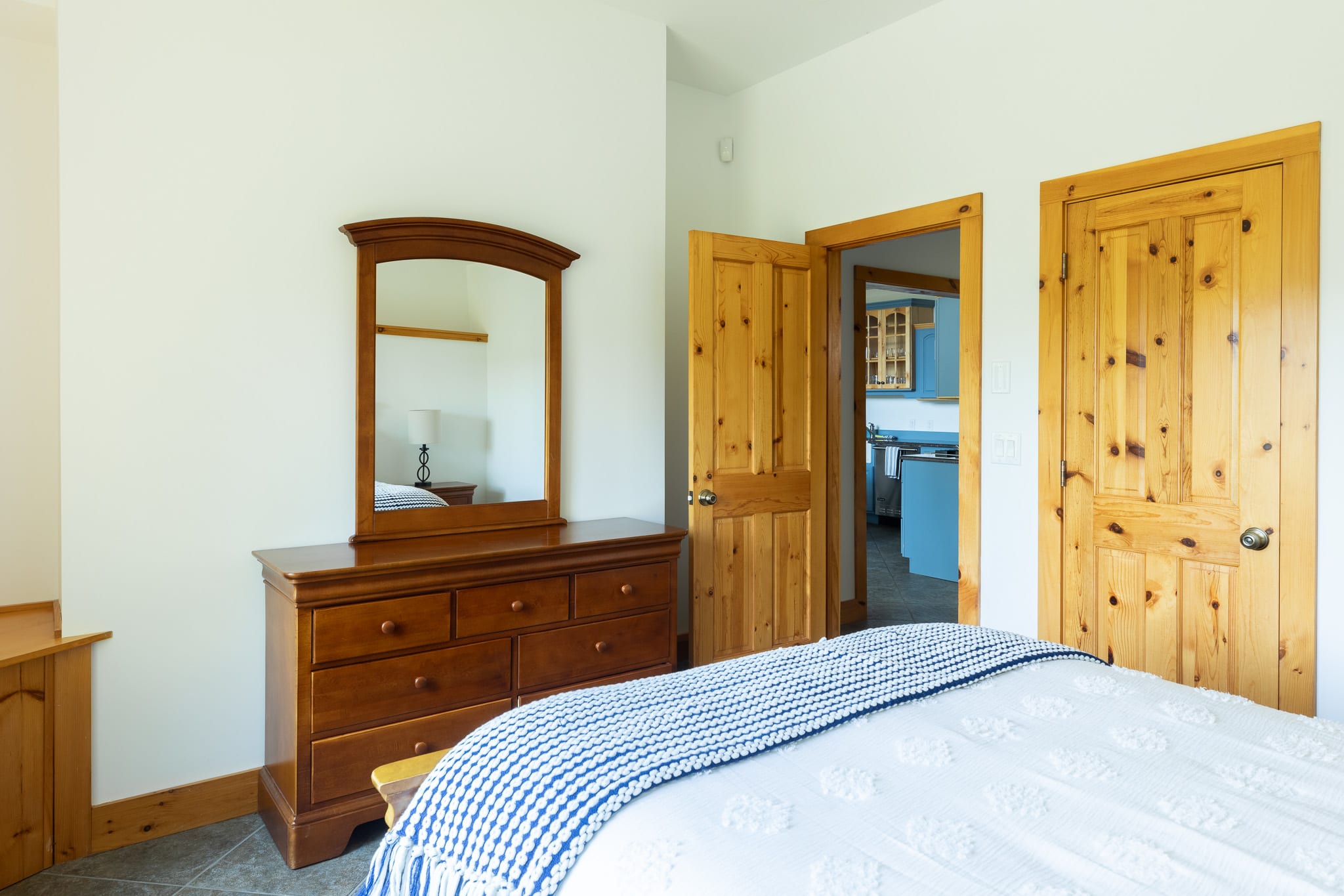 A wooden dresser with an arched mirror stands in a bedroom, reflecting a doorway that opens to a room with blue cabinetry, while a white bed with a blue throw is partially visible.