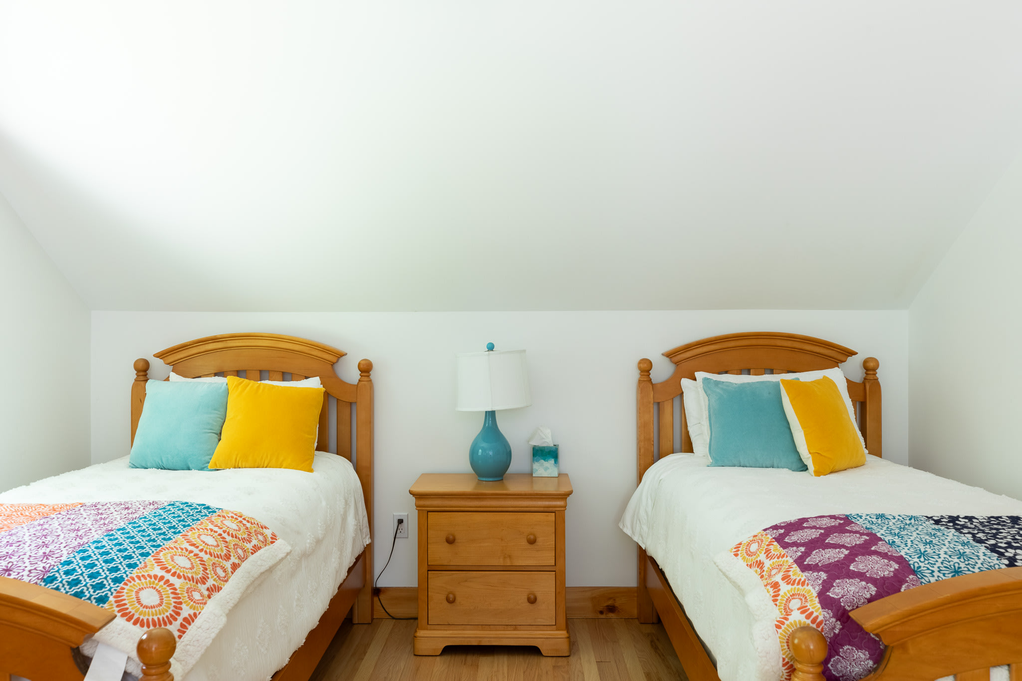 A bright bedroom with two wooden twin beds, each adorned with colorful patterned quilts and blue and yellow throw pillows, separated by a wooden nightstand with a blue lamp.