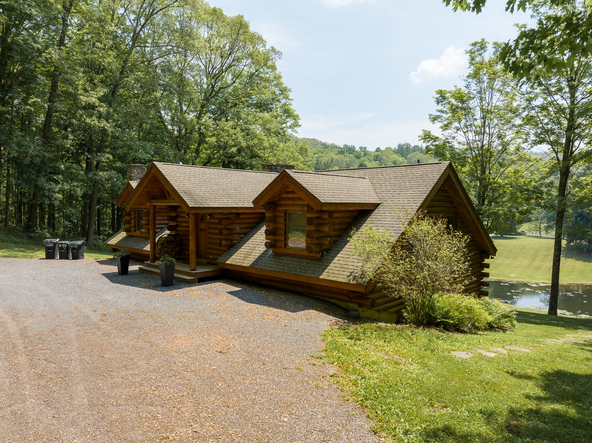 A rustic log cabin with a brown shingle roof and multiple gables, nestled among tall green trees. A gravel driveway leads up to the front entrance, with a small pond visible in the background to the right.