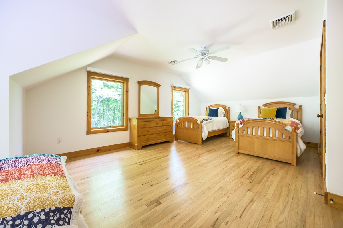 A spacious bedroom featuring two wooden twin beds with colorful quilts, a matching wooden dresser with a mirror, and large windows overlooking lush greenery, all set on polished hardwood floors.