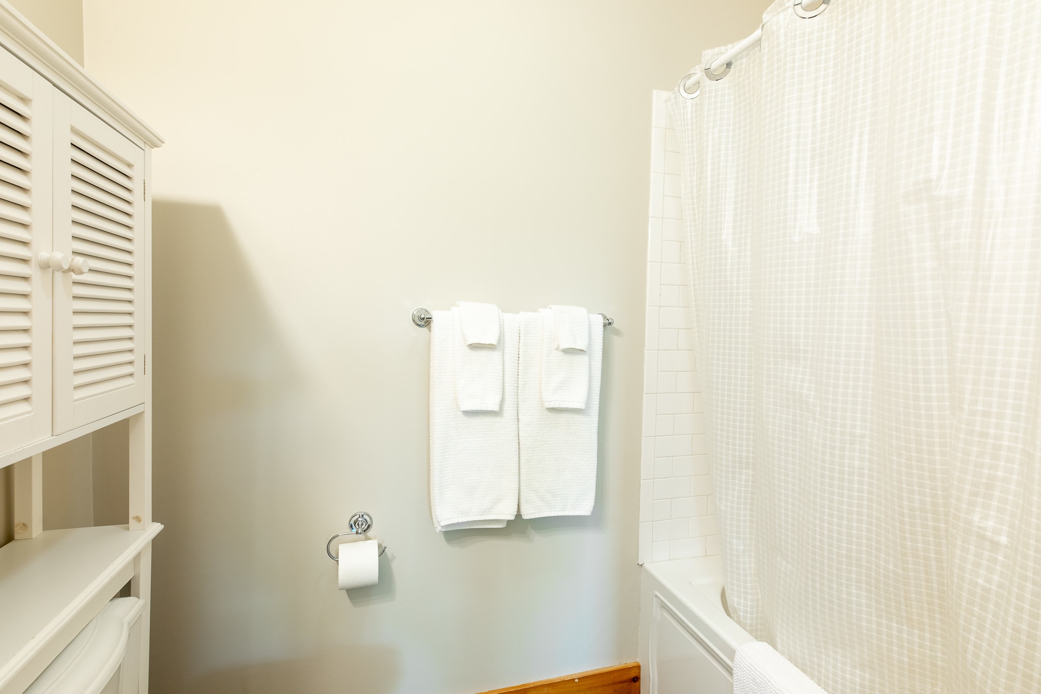 A clean bathroom showing a white toilet with a louvered storage cabinet above it, a towel rack with white towels, and a white shower curtain covering a bathtub.