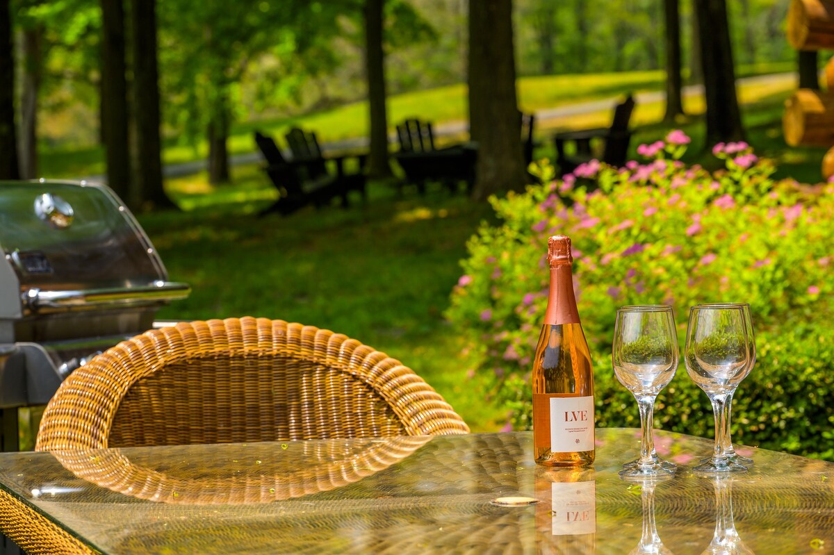 A close-up of an outdoor dining table set with a bottle of rosé wine and two wine glasses, with a BBQ grill and lush greenery in the soft-focus background.