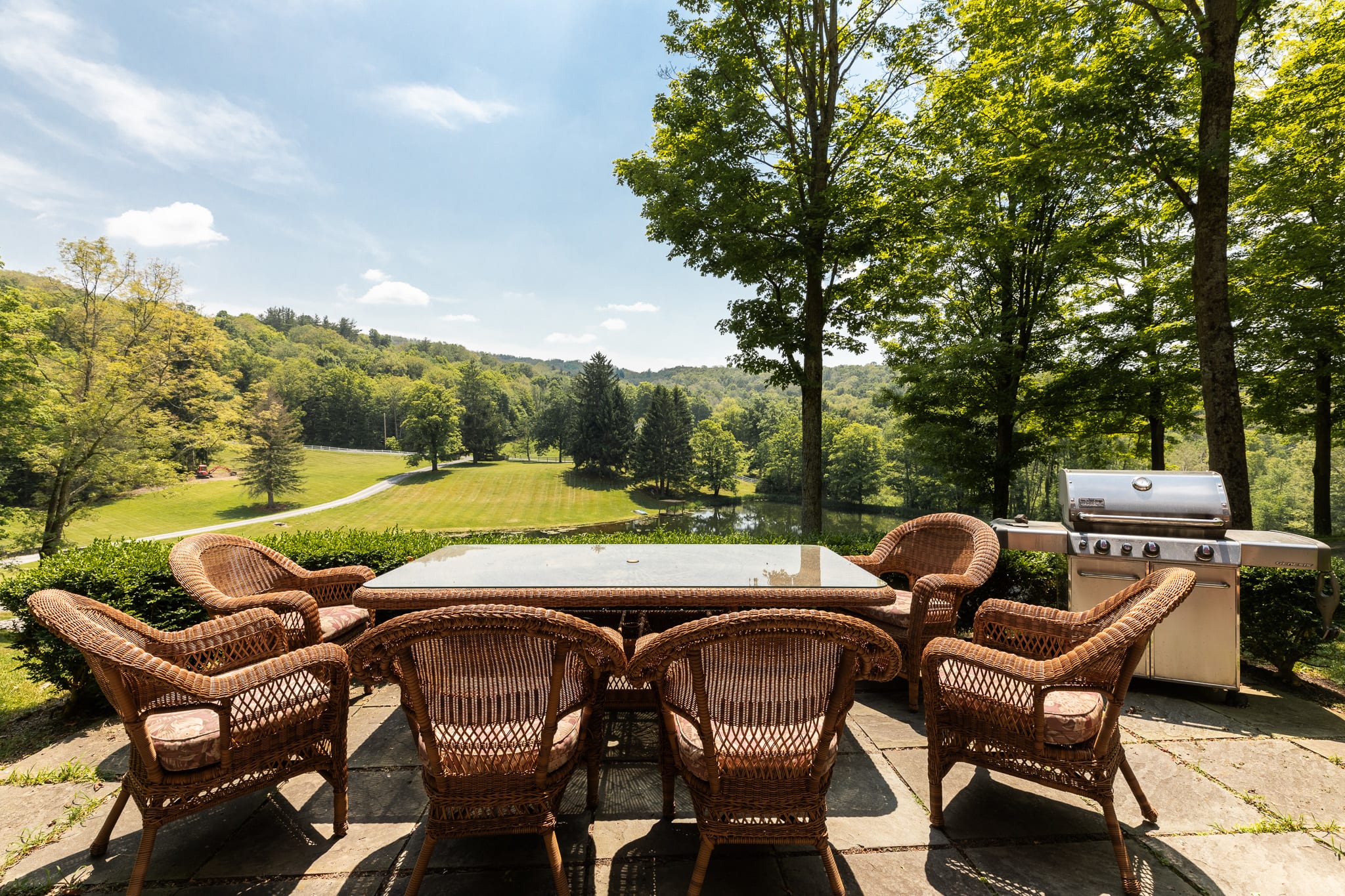 A stone patio with a wicker dining table and chairs, overlooking a scenic valley with a pond and lush green trees under a bright blue sky. A BBQ grill is visible on the side.