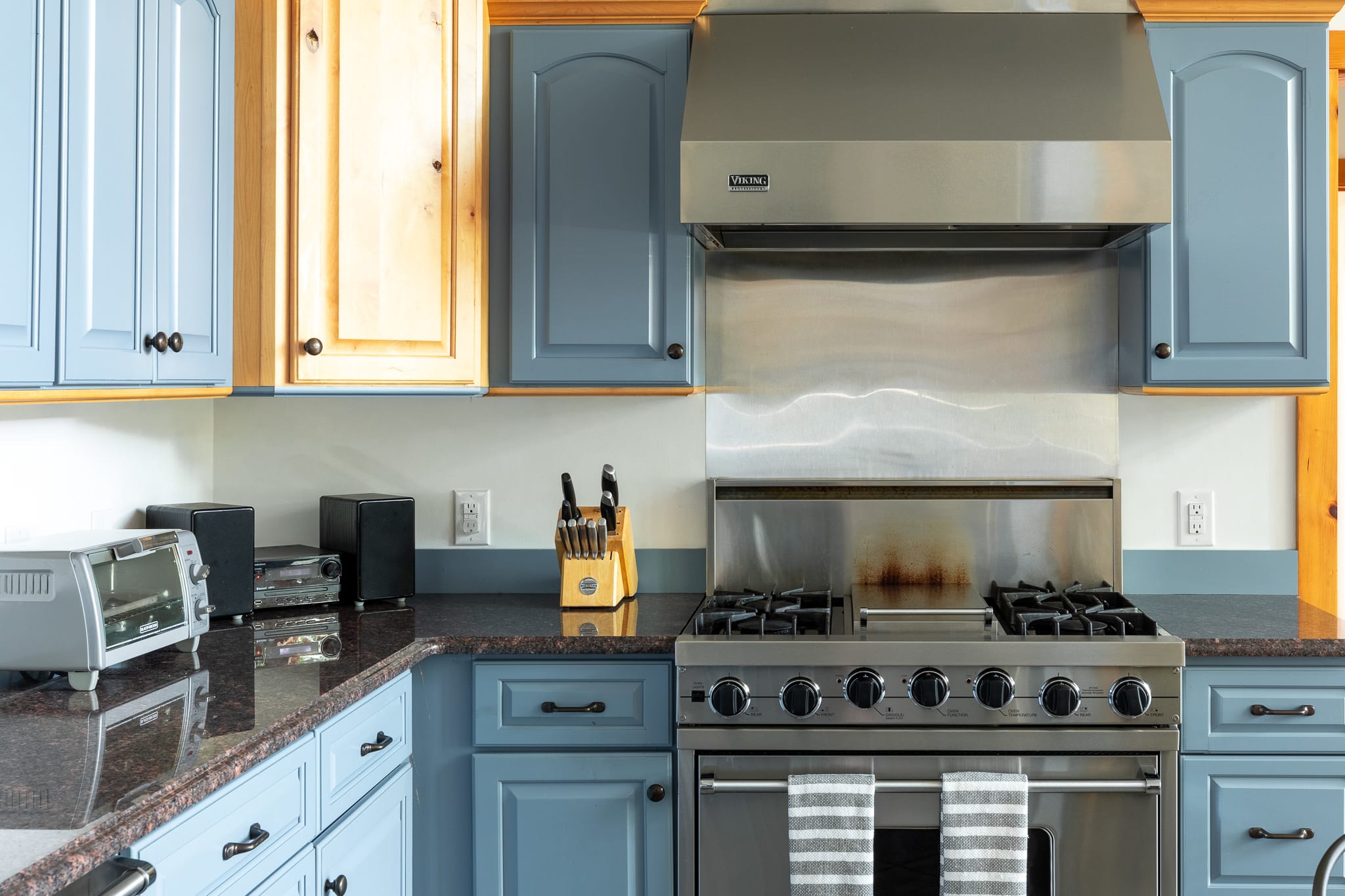 A detailed view of the kitchen counter showcasing a stainless steel gas range with a matching hood, dark granite countertops, and light blue and natural wood cabinetry, with small appliances visible.