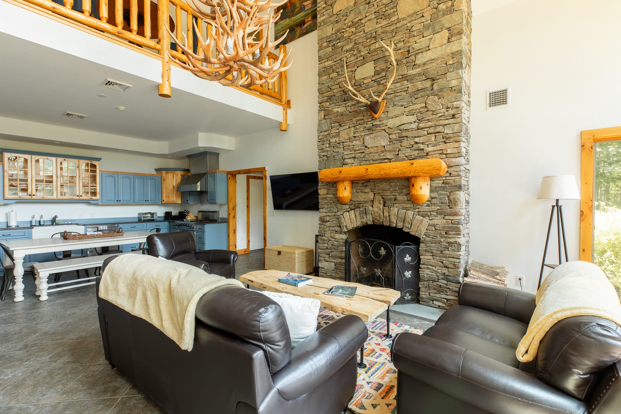A wide shot of the living room features a prominent stone fireplace with a wooden mantel, dark leather seating, and a view into the adjacent kitchen area with blue cabinets. A large antler chandelier hangs above.