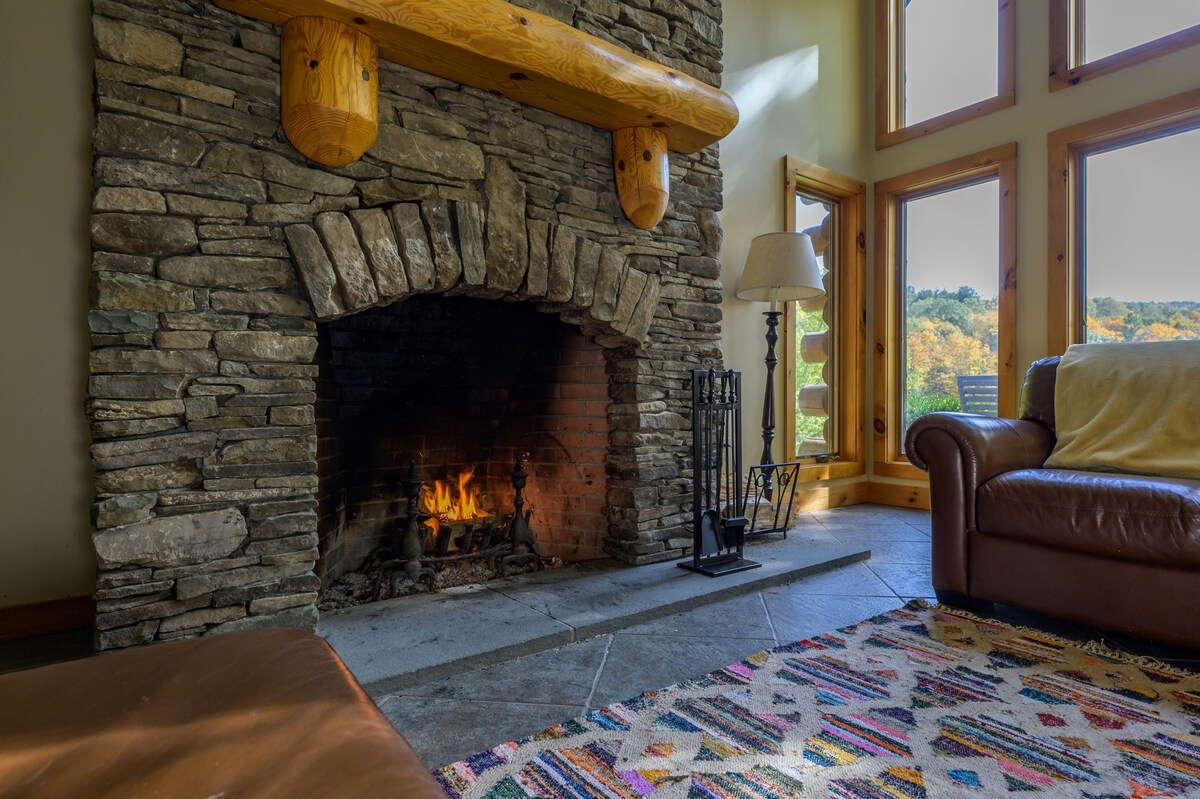 A close-up of the rustic stone fireplace with a roaring fire, a heavy wooden mantel, and a black metal fire screen, flanked by a floor lamp and a brown leather armchair, all on a tiled floor.