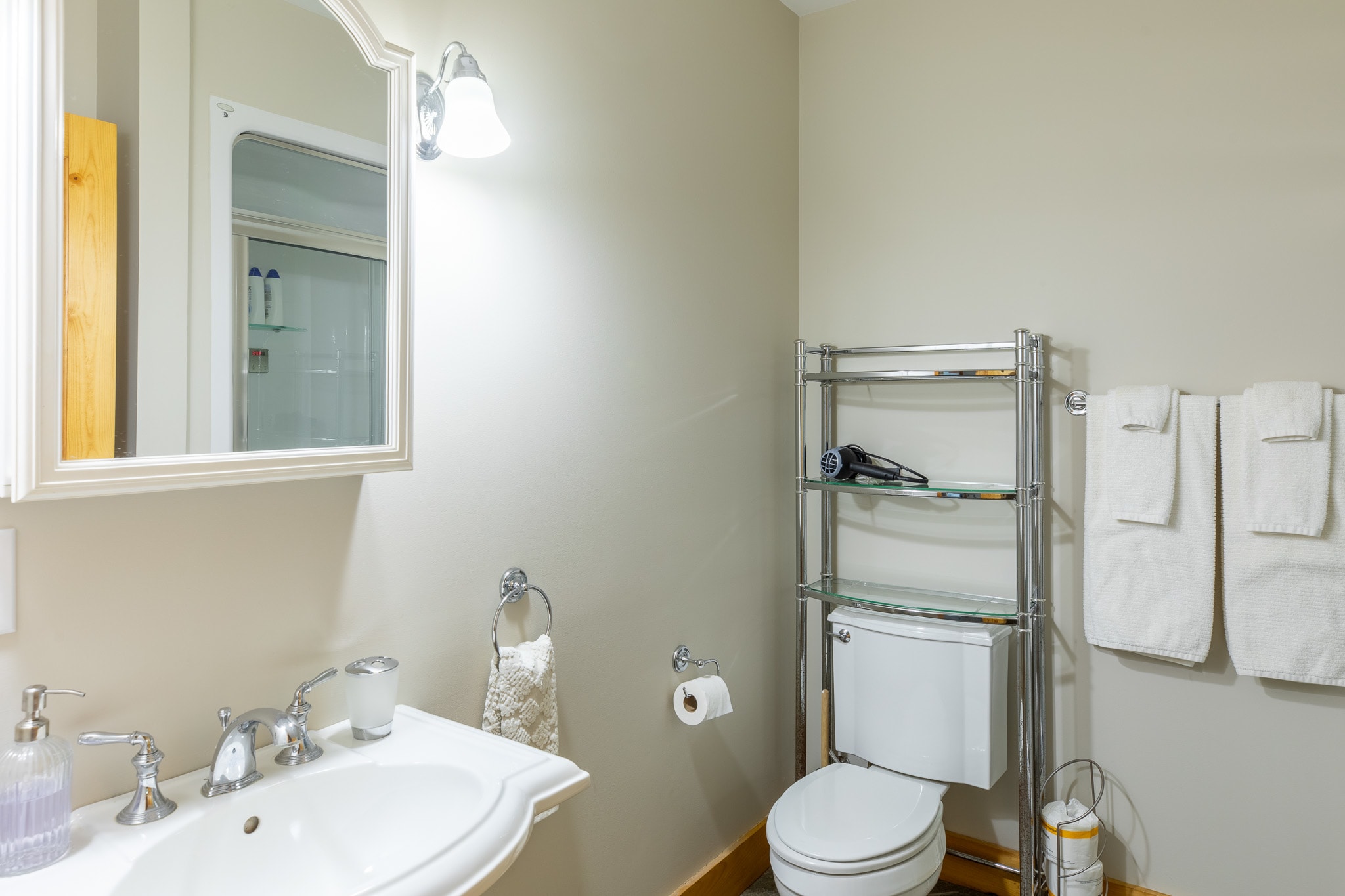 A clean bathroom featuring a white pedestal sink with a chrome faucet, a mirrored medicine cabinet, a toilet with a chrome shelving unit above it, and light beige walls.
