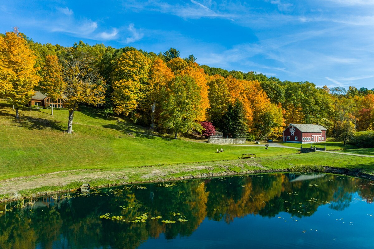 A vibrant autumn landscape featuring a serene pond reflecting the blue sky, surrounded by lush green grass and a hillside covered in trees with brilliant yellow and orange fall foliage. A red barn is visible on the right, and a log cabin is nestled among the trees on the left.