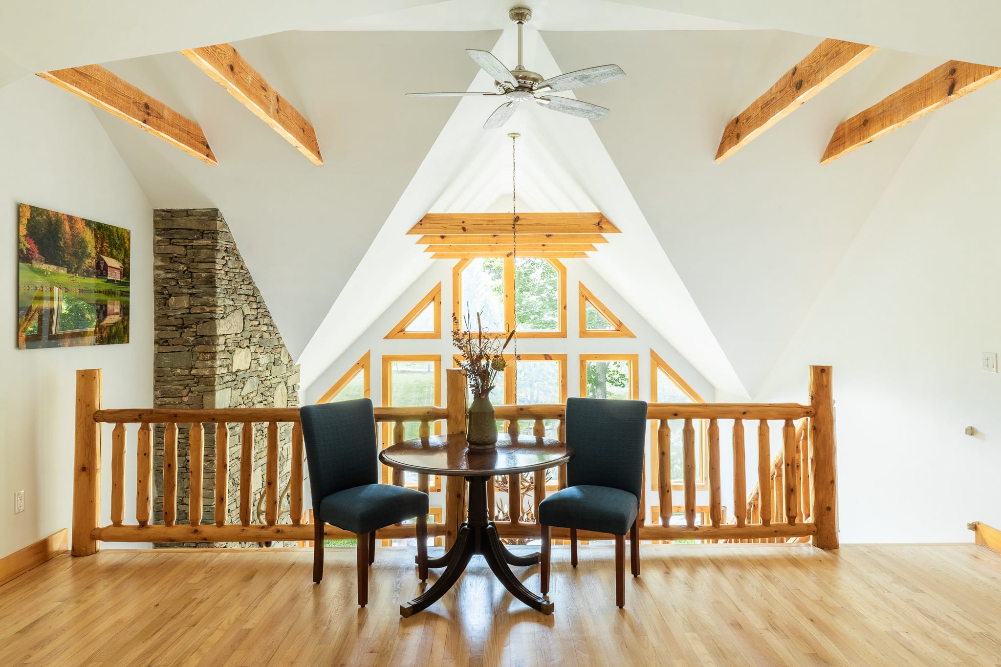 A bright loft area overlooking the living space, featuring a small round wooden table with two blue chairs, exposed wooden beams, and a ceiling fan, with large windows providing natural light and hardwood floors.