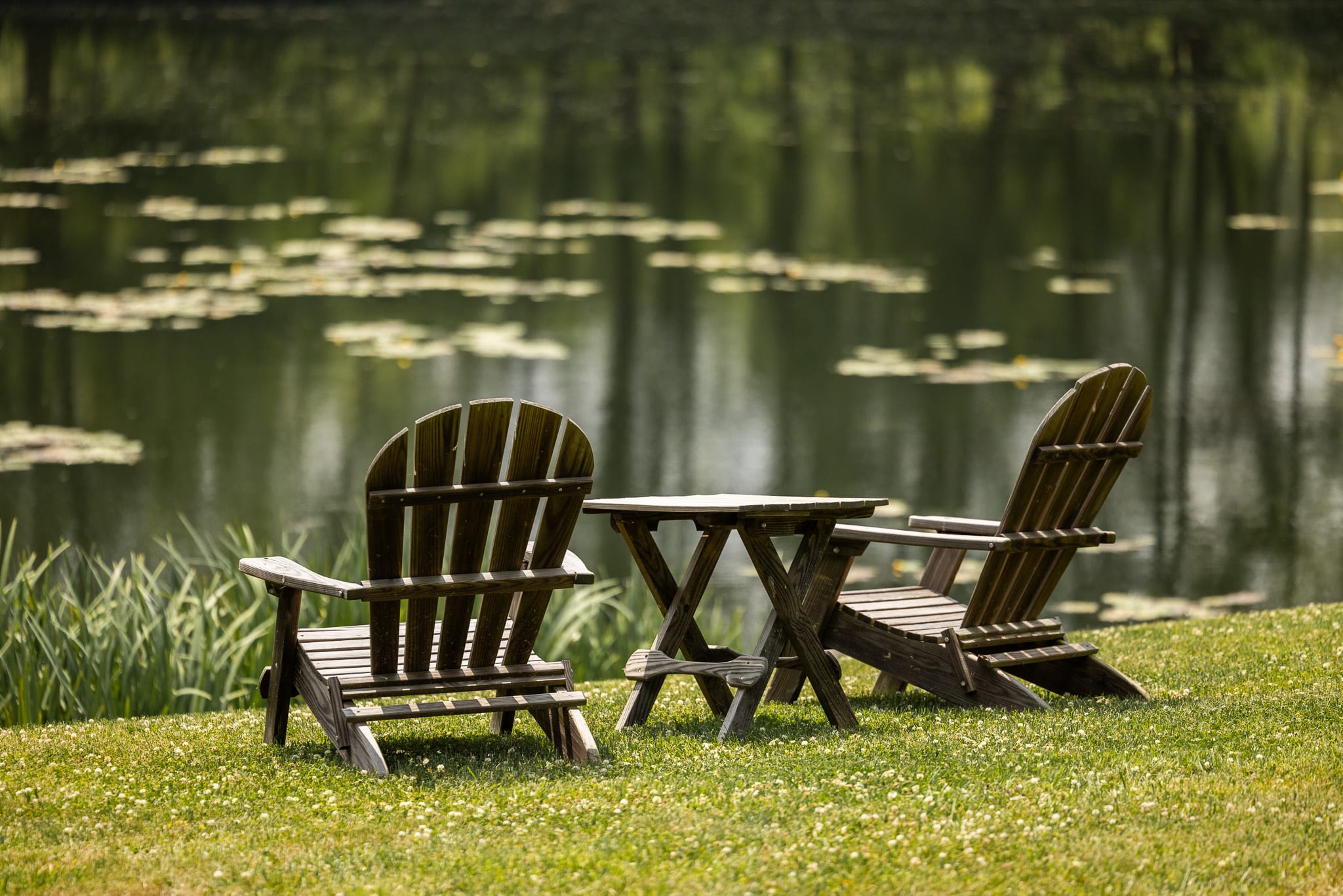 Two wooden Adirondack chairs and a small matching table are set on a grassy lawn beside a serene pond with reflections and lily pads.