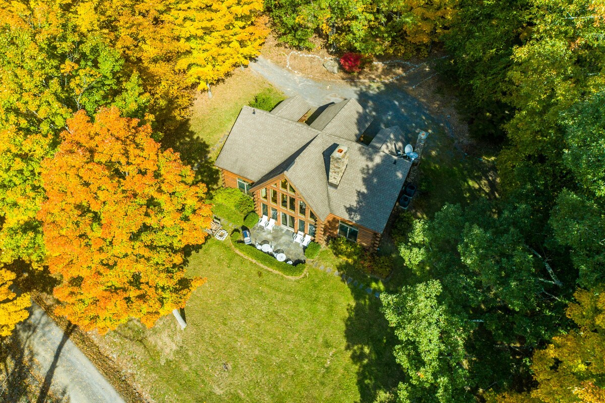 An aerial view of a charming log cabin with a large A-frame window section, surrounded by a vibrant canopy of yellow and orange autumn trees. An outdoor seating area with white chairs is visible on the green lawn beside the cabin.