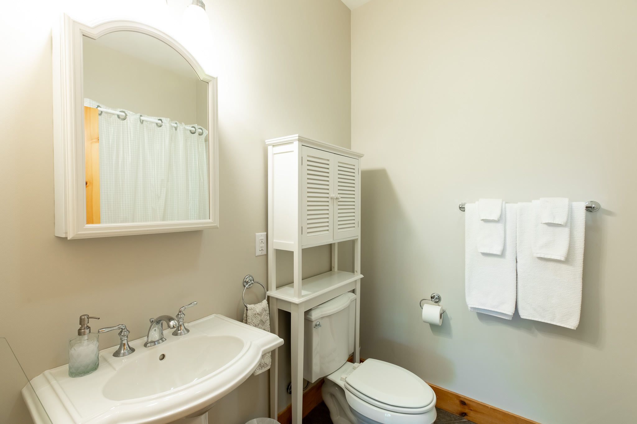 A bright bathroom with light beige walls, a white pedestal sink with a chrome faucet, a white toilet, and a white cabinet with louvered doors above the toilet. An arched mirror hangs above the sink.