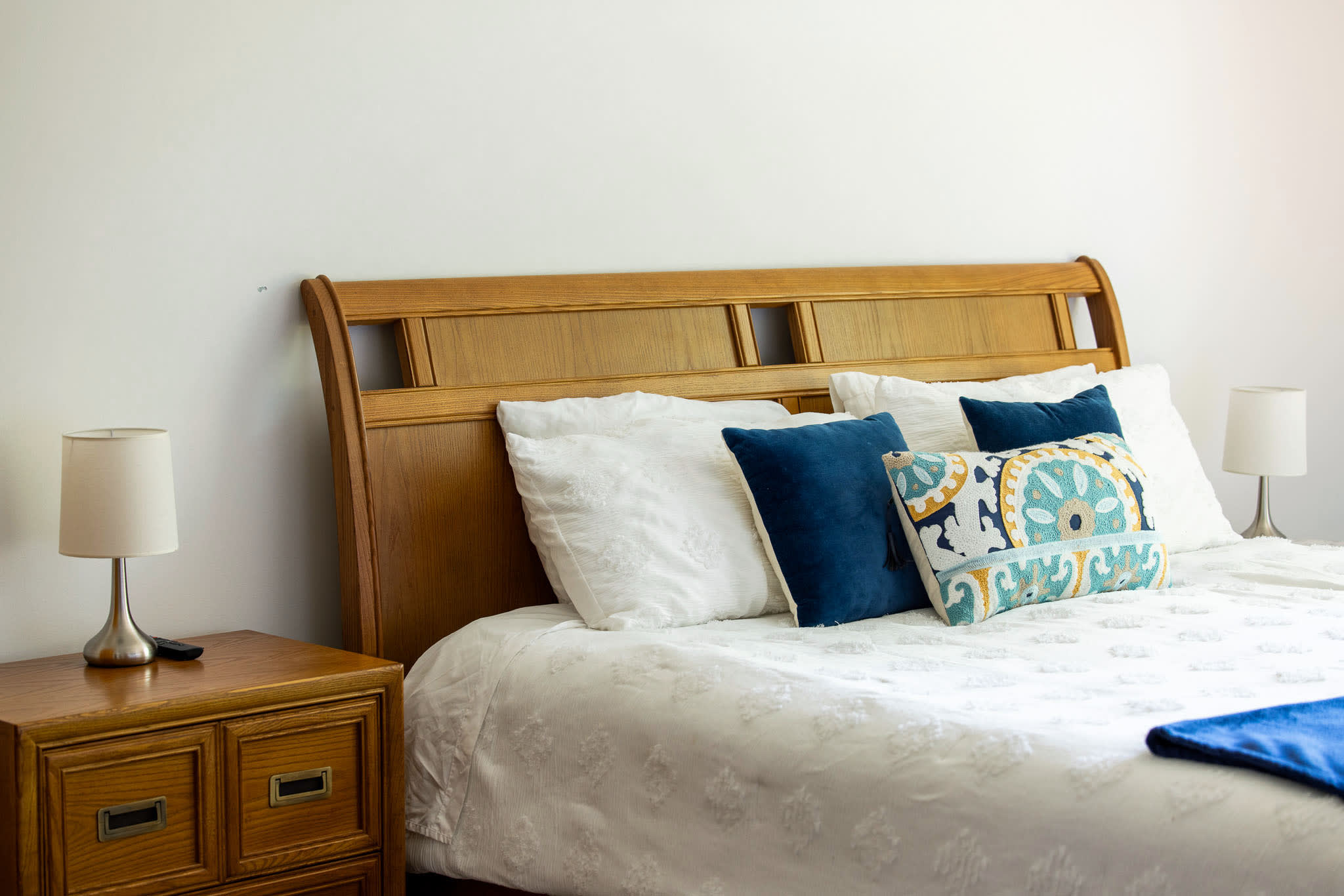 A cozy bedroom featuring a wooden sleigh bed with white bedding, accented by blue and patterned throw pillows, flanked by wooden nightstands with white lamps.