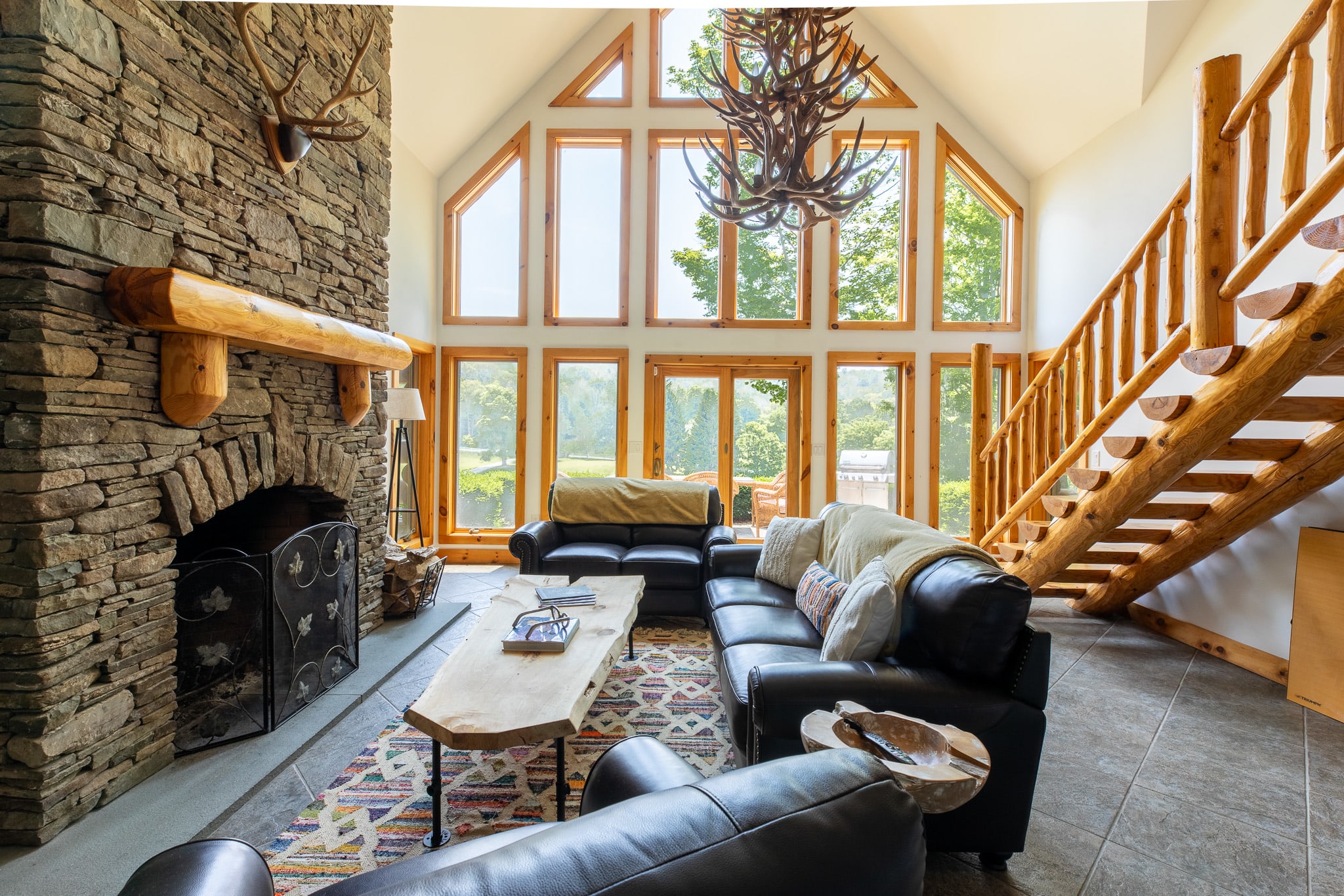 A spacious living room features a large stone fireplace, two dark leather sofas, and a unique live-edge wood coffee table, all set against a backdrop of tall windows offering views of lush greenery. A large antler chandelier hangs from the vaulted ceiling.