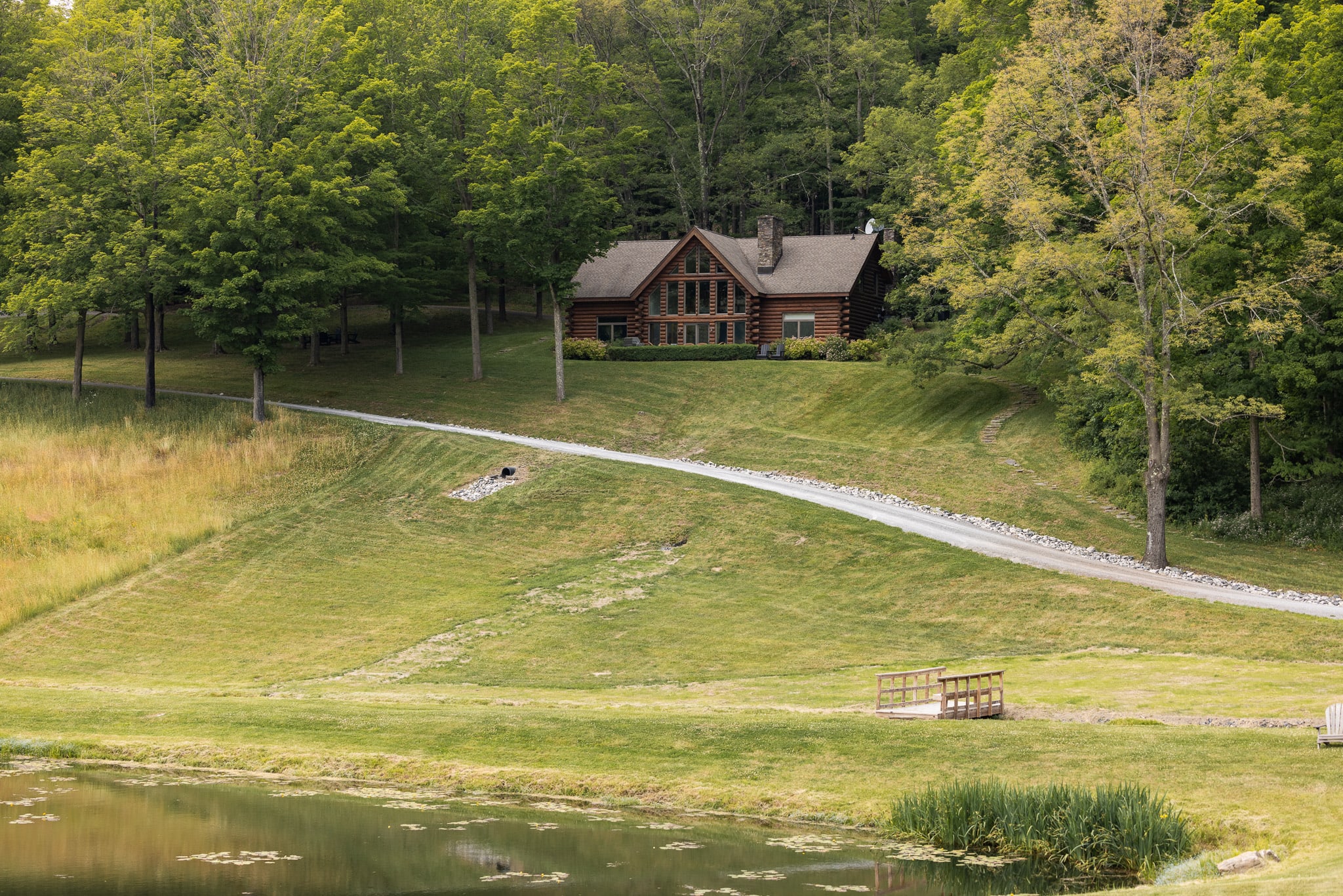 A picturesque log cabin sits atop a grassy hill, accessed by a winding gravel driveway, overlooking a tranquil pond with a small wooden dock. The landscape is dotted with mature green trees under a bright sky.