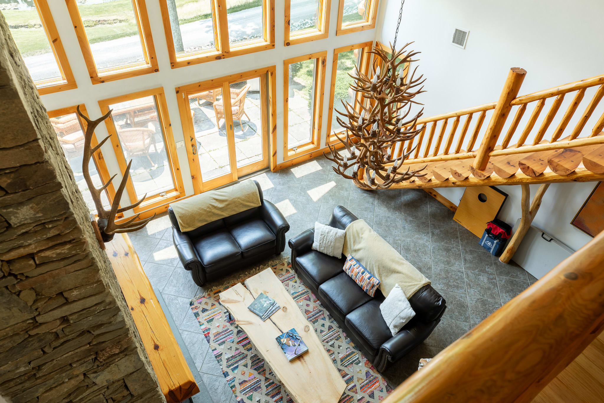 An elevated view of the spacious living room, showcasing the high ceilings, large windows, a stone fireplace, and a comfortable seating area with leather furniture, all beneath a grand antler chandelier.