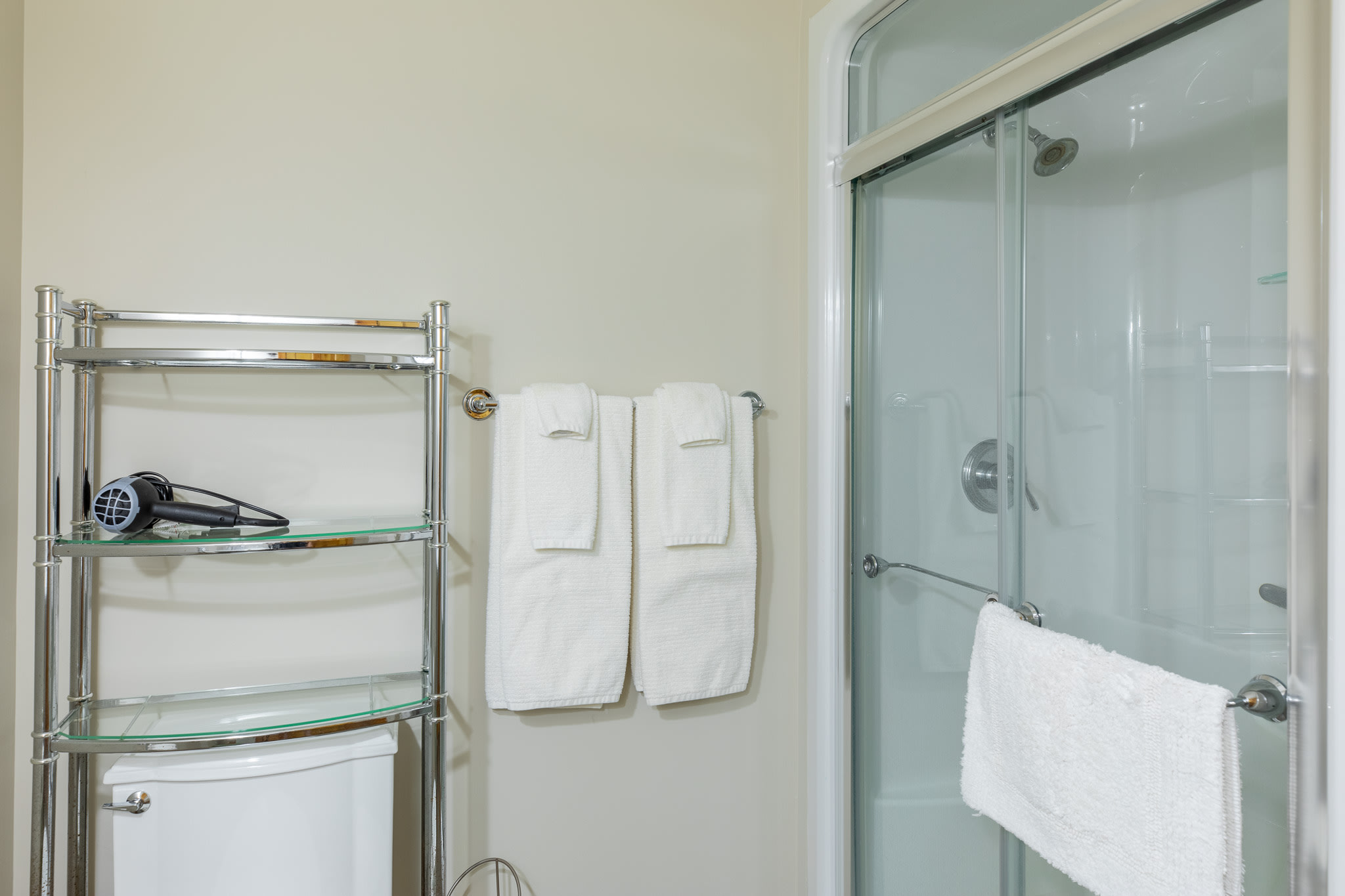 A bright bathroom showcasing a white toilet with a chrome shelving unit holding a hairdryer, fresh white towels hanging on the wall, and a glass-enclosed shower stall.