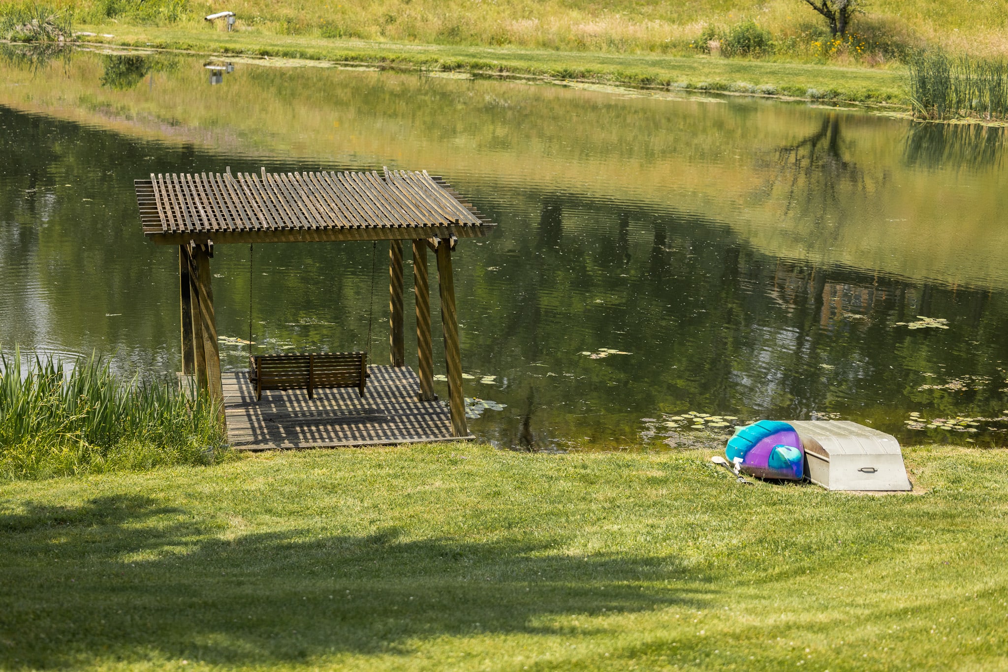 A wooden dock with a swing bench, situated on the grassy bank of a calm pond with lily pads, next to two small boats.