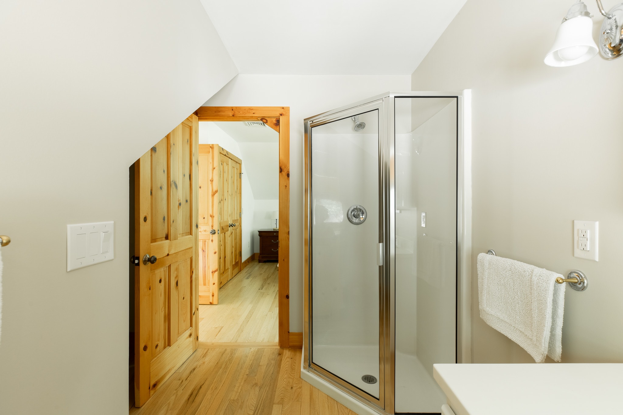 A different angle of the bathroom, showing the glass-enclosed shower and the white vanity, with a view into the adjacent bedroom through an open wooden door, all on hardwood floors.
