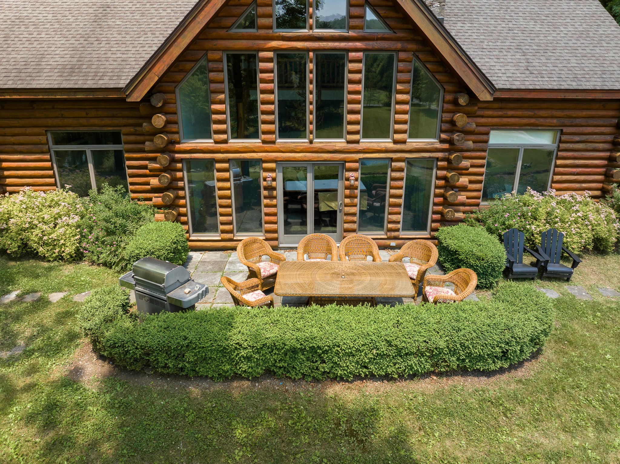 An aerial view of the back of a large log cabin with a stone patio featuring a wicker dining set and a BBQ grill, surrounded by manicured hedges and green lawn.