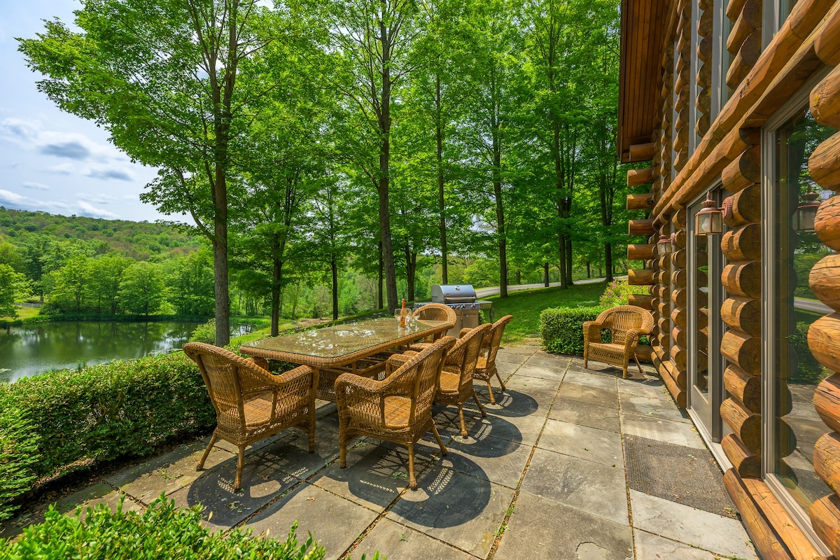 A stone patio with a wicker dining set and a BBQ grill, offering views of a tranquil pond and surrounded by lush green trees and manicured bushes.