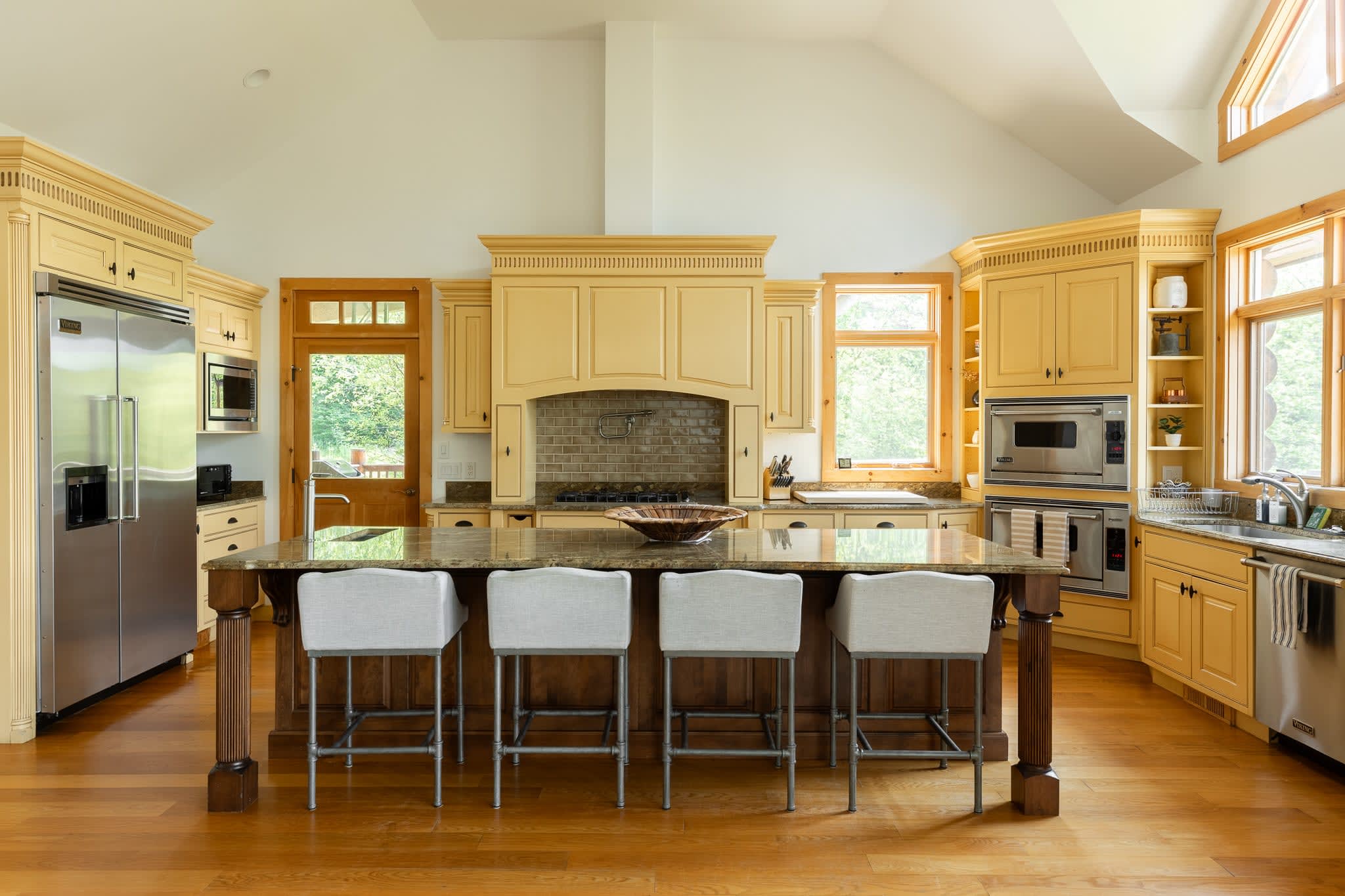 Well-lit kitchen featuring a large wooden island with four bar stools, set against light yellow cabinets and granite countertops. The gas range and built-in ovens are visible, with natural light streaming through the windows.