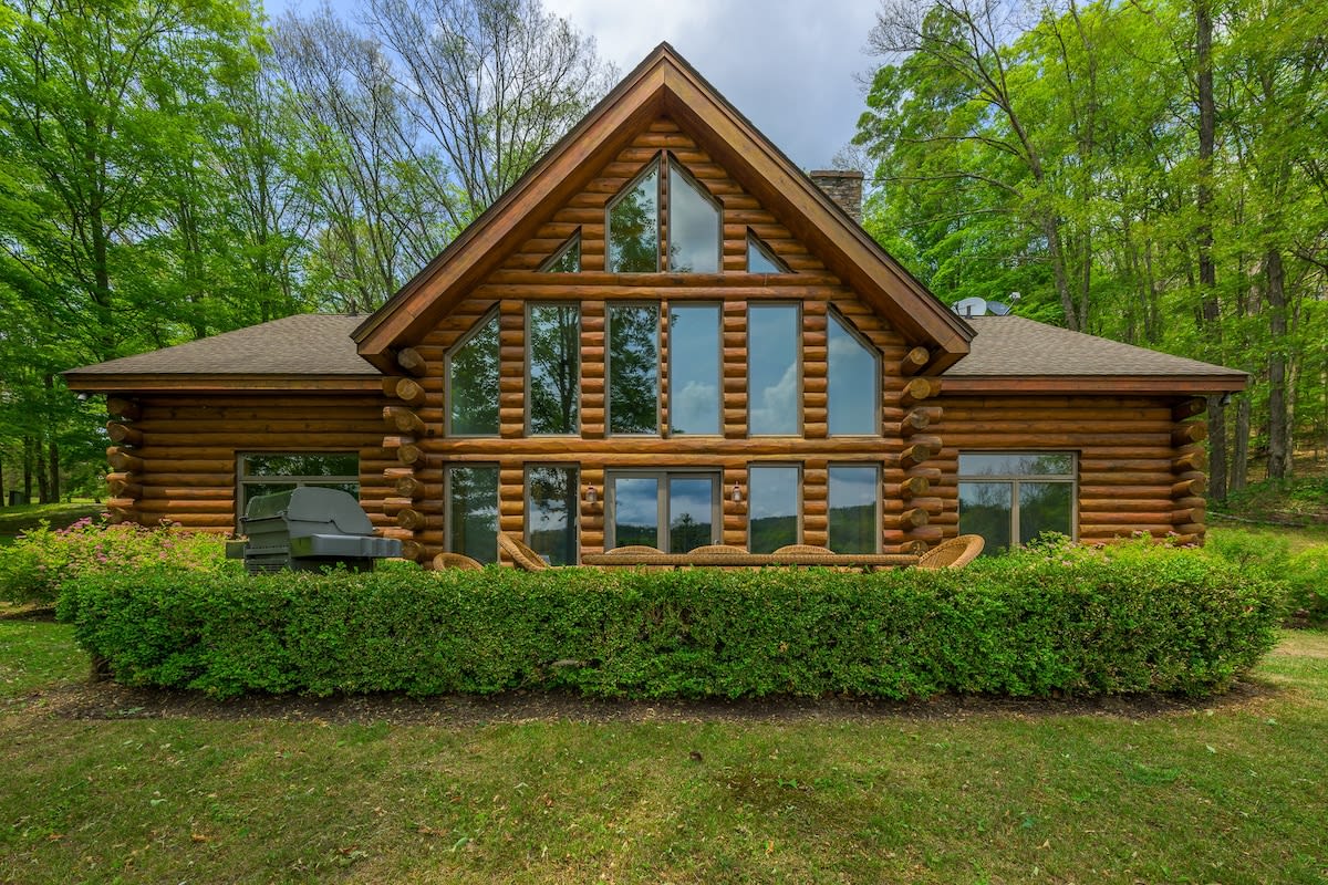 Full exterior view of a rustic log cabin with a prominent A-frame and large windows, set amidst a lush green forest. A hedge lines the front of the house, with a grill and outdoor seating visible.
