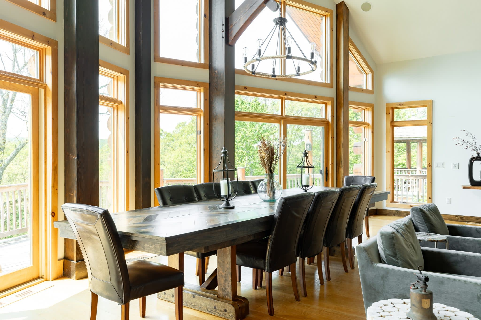 A long, dark wood dining table with a distinctive herringbone pattern top is surrounded by dark leather chairs, set against a backdrop of expansive floor-to-ceiling windows that offer bright natural light and views of lush greenery. A large chandelier hangs overhead.