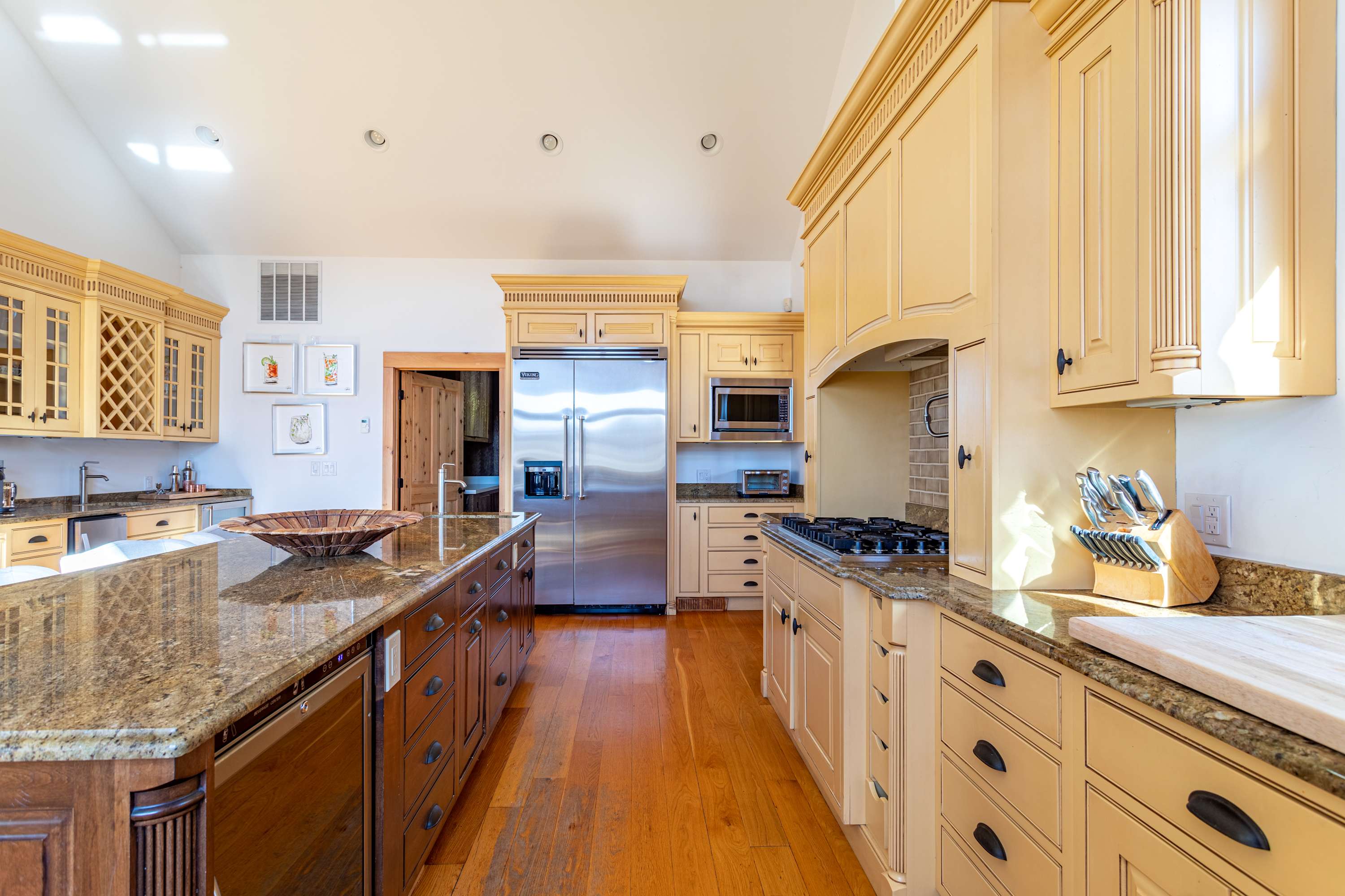 Spacious kitchen featuring a large central island with dark wood base and granite countertop, surrounded by light yellow cabinetry. A large stainless steel refrigerator is prominent, and hardwood floors run throughout the space.