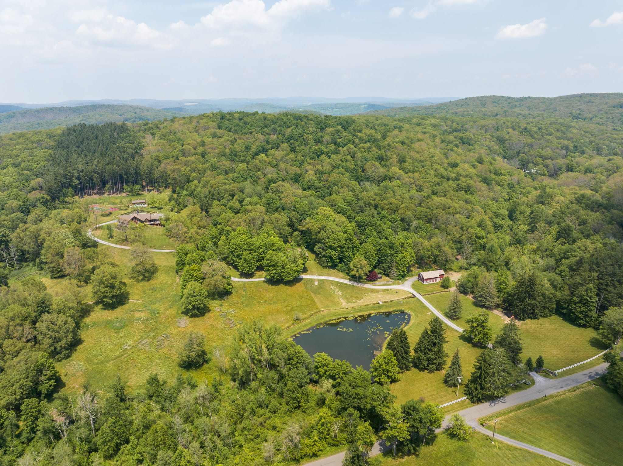 A wide aerial shot revealing the expansive property, featuring multiple log cabin structures, a serene pond, and winding driveways, all set amidst rolling green hills and dense forests under a clear sky.