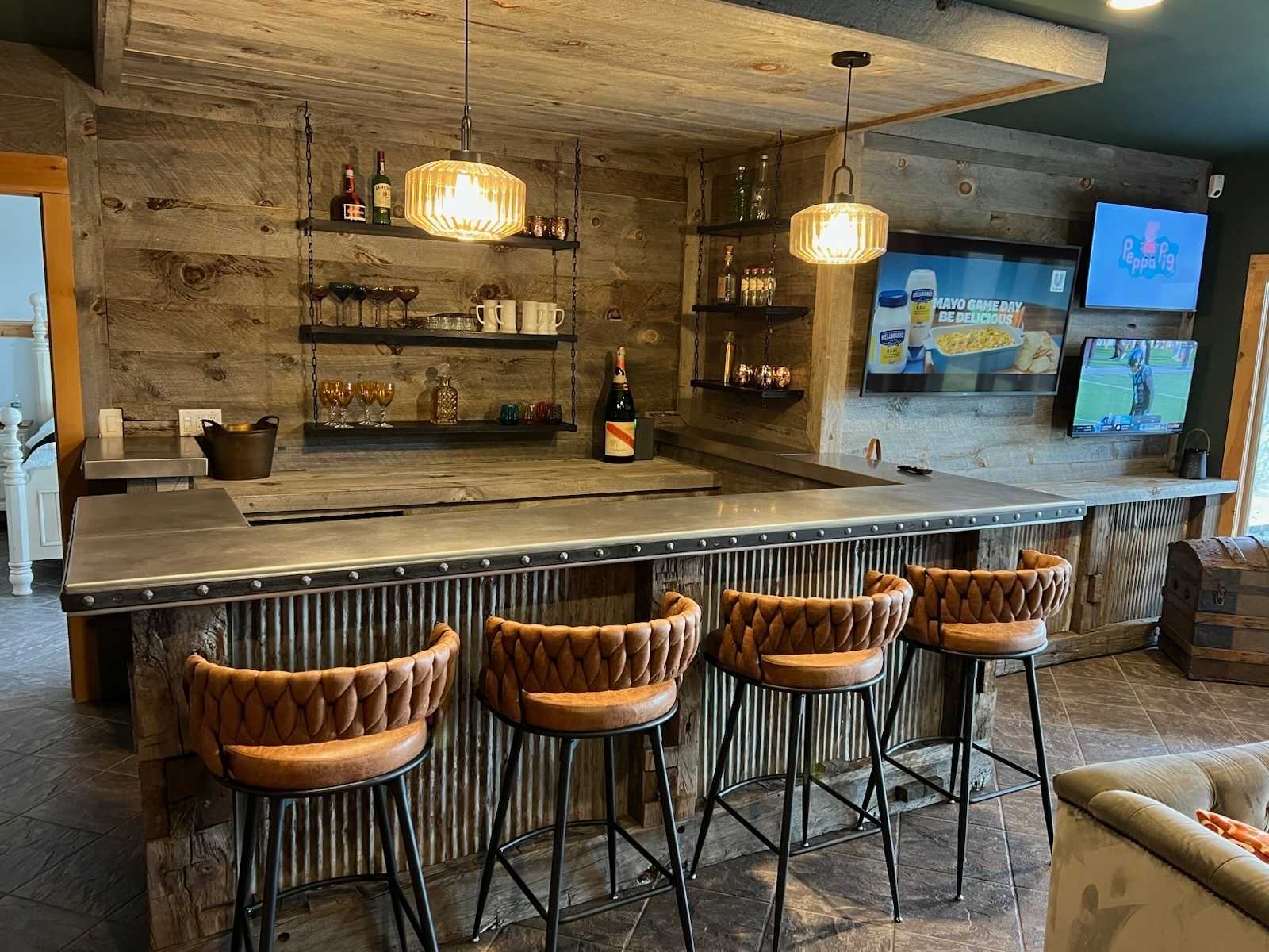 A detailed view of the rustic wooden bar area, featuring a corrugated metal front, a metal countertop, and four brown leather-braided bar stools. Shelves with bottles and glasses are mounted on the reclaimed wood wall, illuminated by two pendant lights.