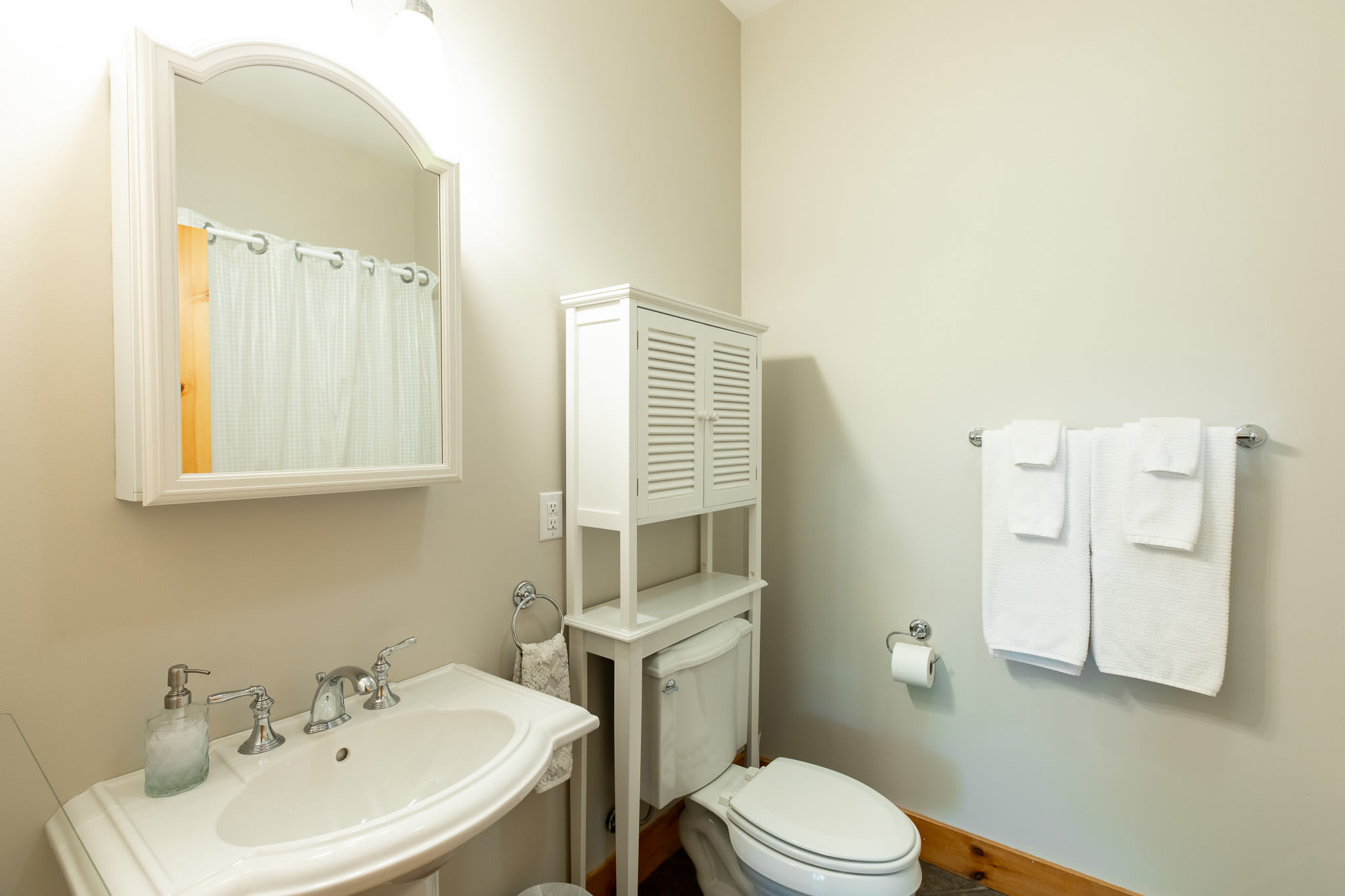 A light-colored bathroom features a white pedestal sink, a decorative arched mirror above, and a white wooden cabinet over the toilet, with white towels on a rack.