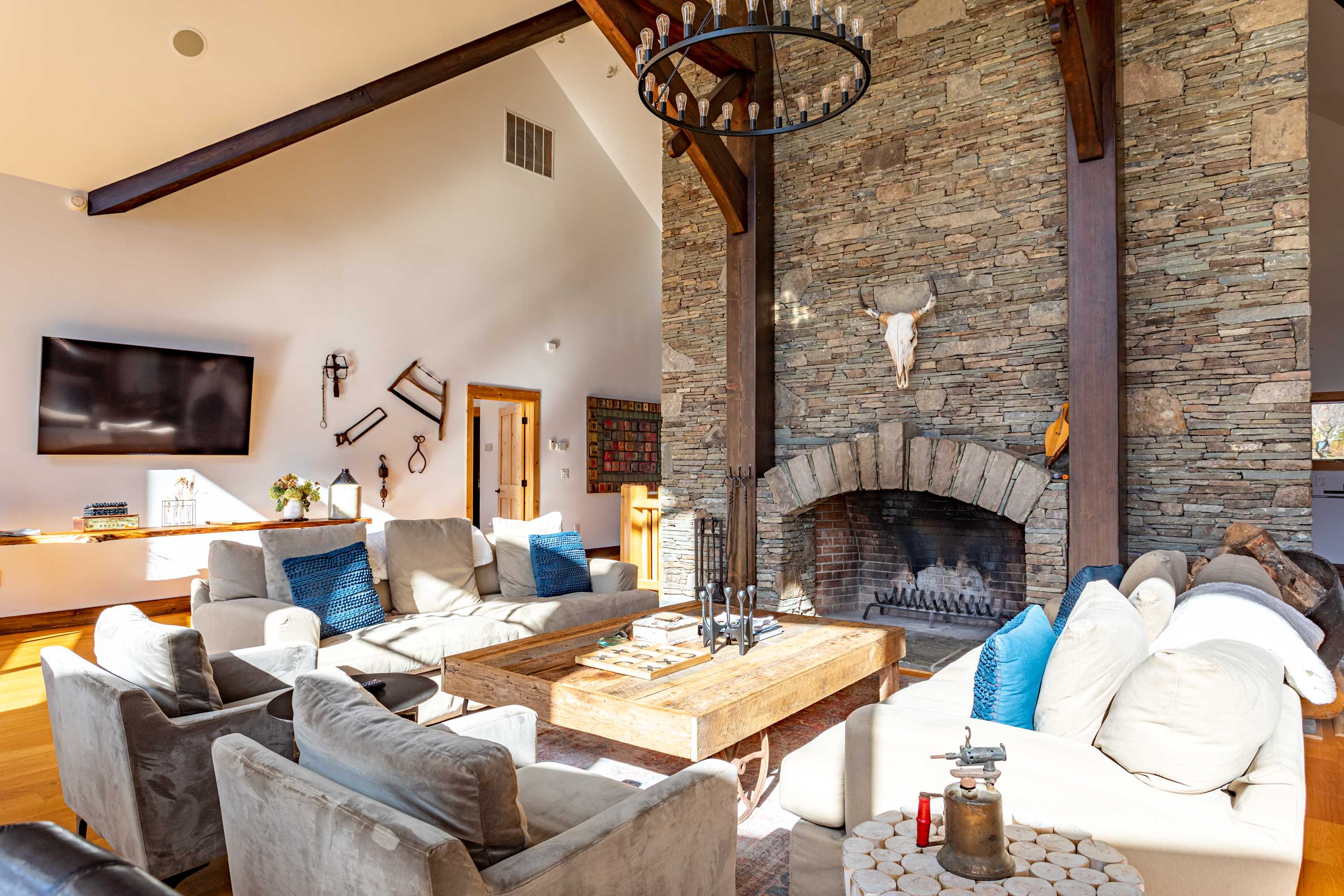 A spacious living room features multiple light-colored sofas and armchairs arranged around a large rustic wooden coffee table, all facing a grand stone fireplace with an animal skull. Exposed wooden beams and a high vaulted ceiling add to the rustic charm.