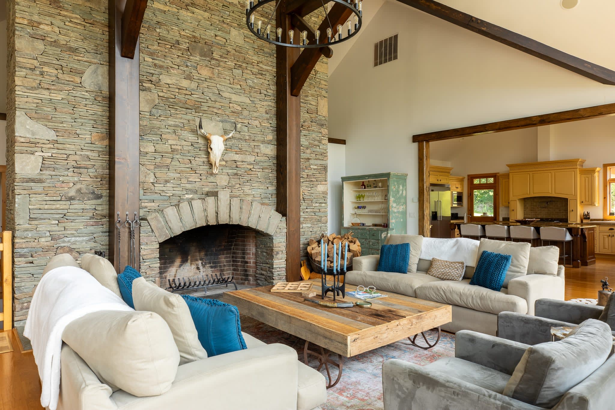 This inviting living room centers around a large stone fireplace adorned with an animal skull, surrounded by comfortable sofas and armchairs with blue accent pillows. A rustic wooden coffee table and a distressed green cabinet add character, with the kitchen visible in the background.