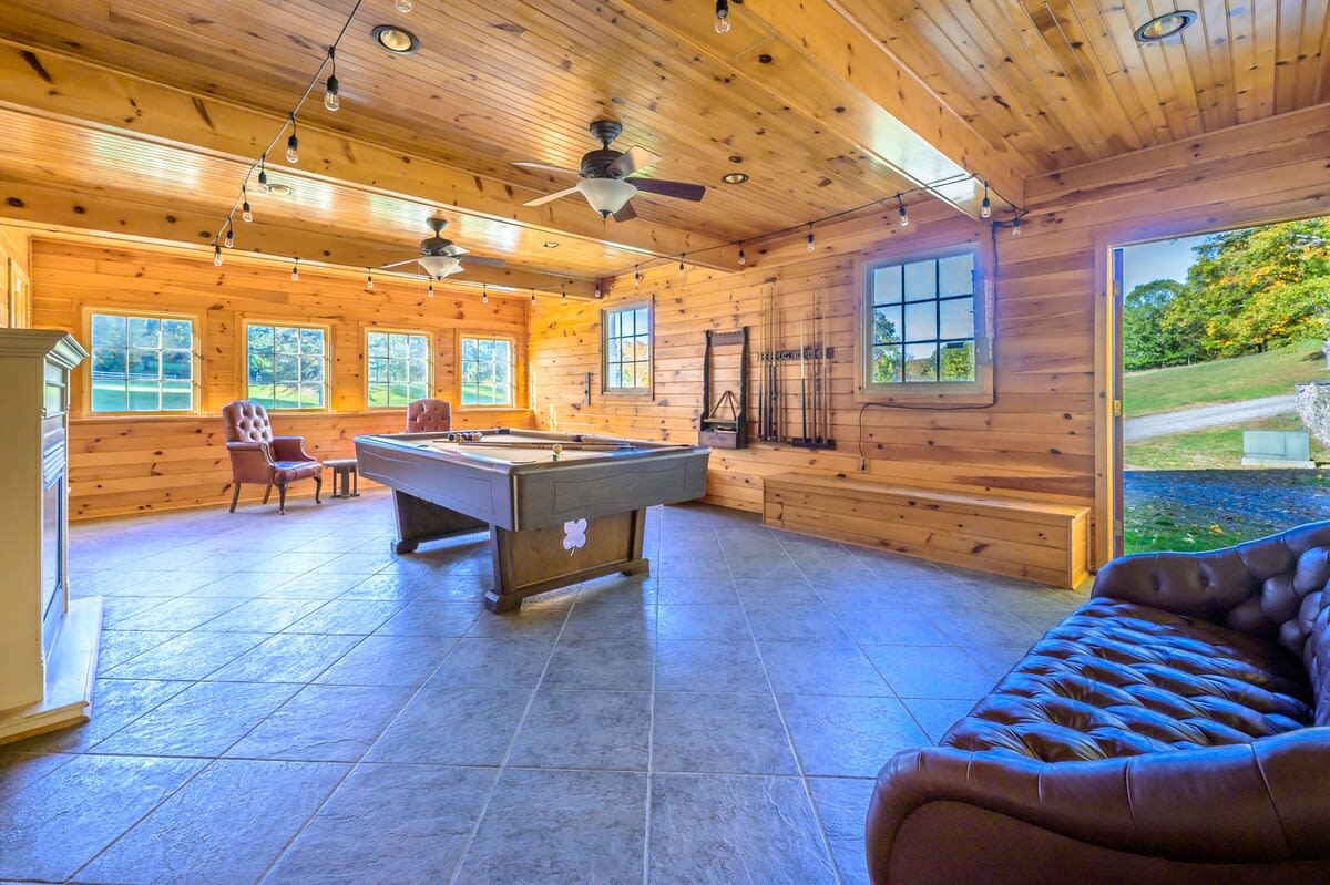 A rustic game room features a pool table in the center, surrounded by warm wooden paneled walls and ceiling, with multiple windows letting in natural light.