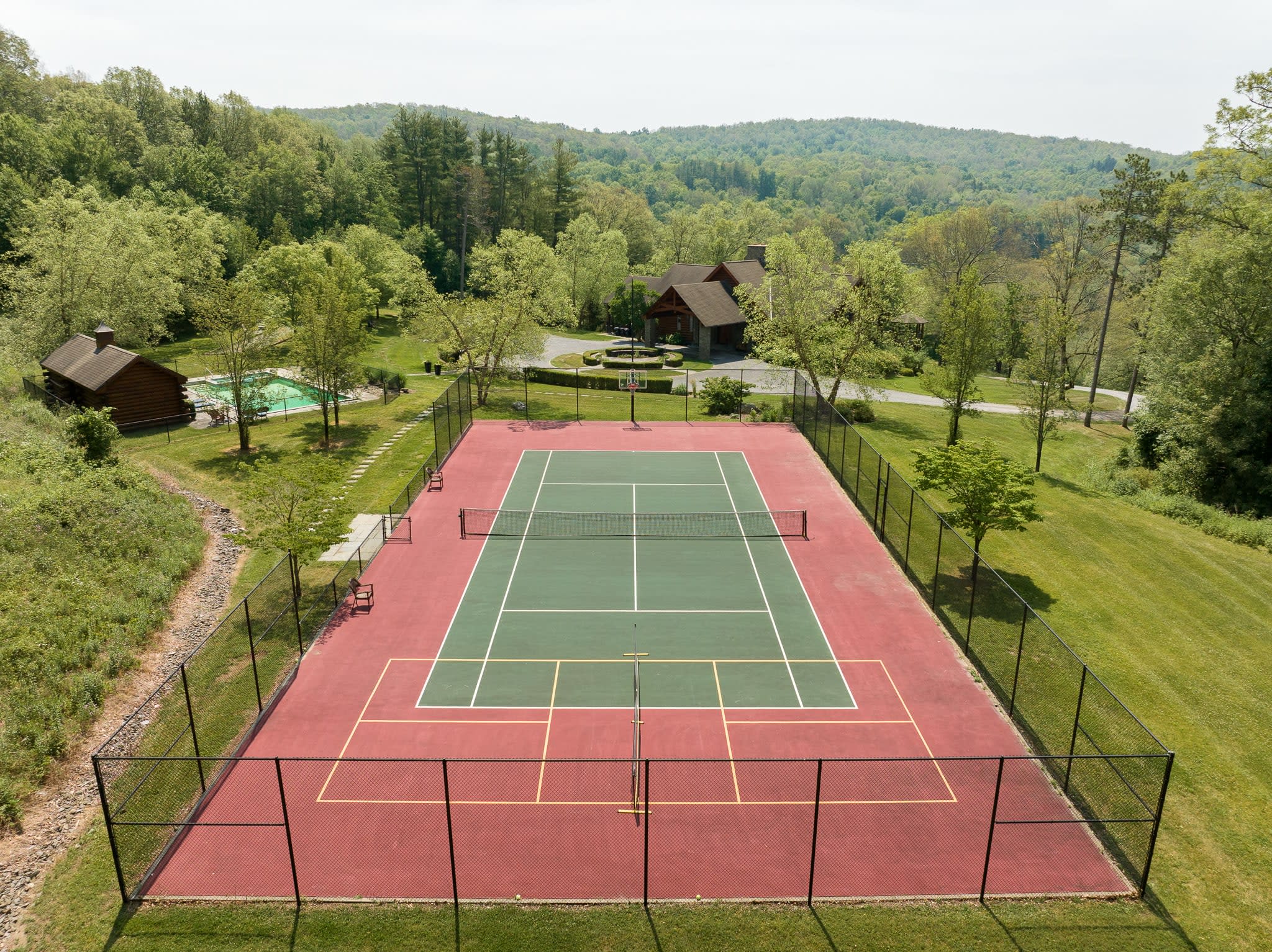 Aerial view of a red and green fenced tennis court, with a pool and the main lodge visible in the background, surrounded by dense green forests and rolling hills.