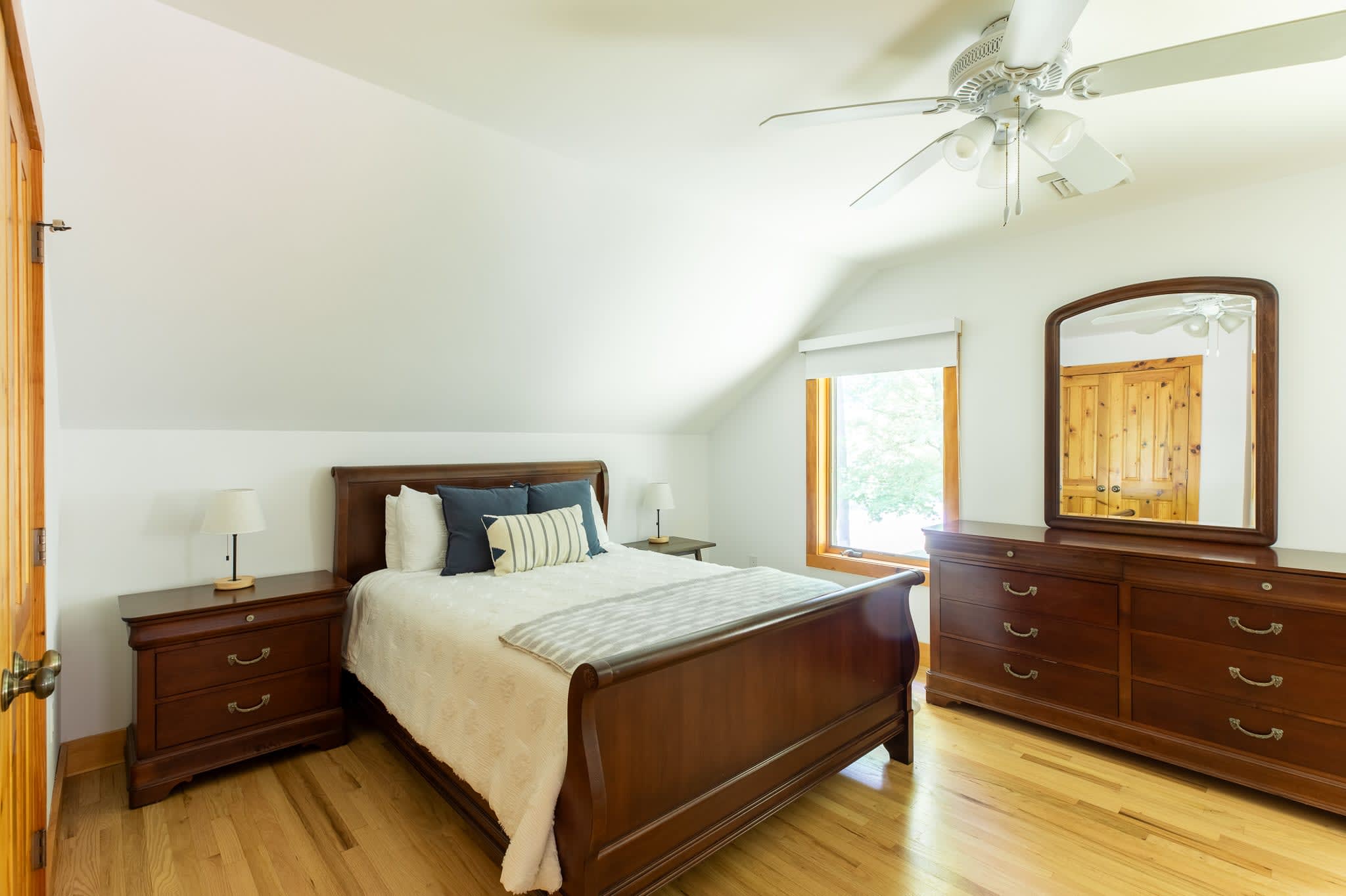 Bedroom-1 features a dark wood sleigh bed with white bedding and blue accent pillows, flanked by matching nightstands with lamps, and a large dresser with a mirror, all on light hardwood floors under a ceiling fan.