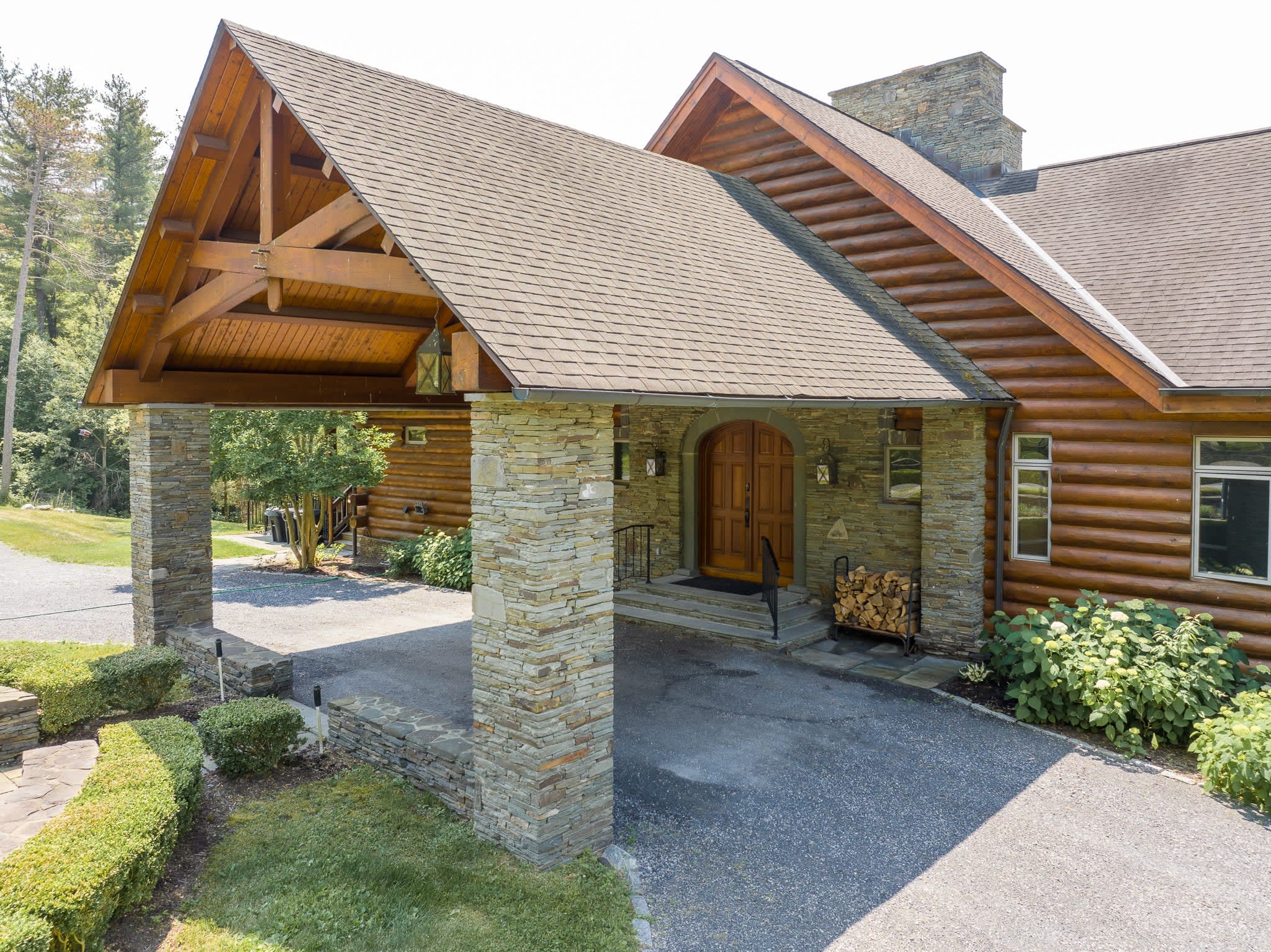 Front exterior of a log cabin featuring a grand covered entrance with stone pillars and exposed wooden beams. Double wooden doors lead into the home, with neatly stacked firewood beside the entrance.