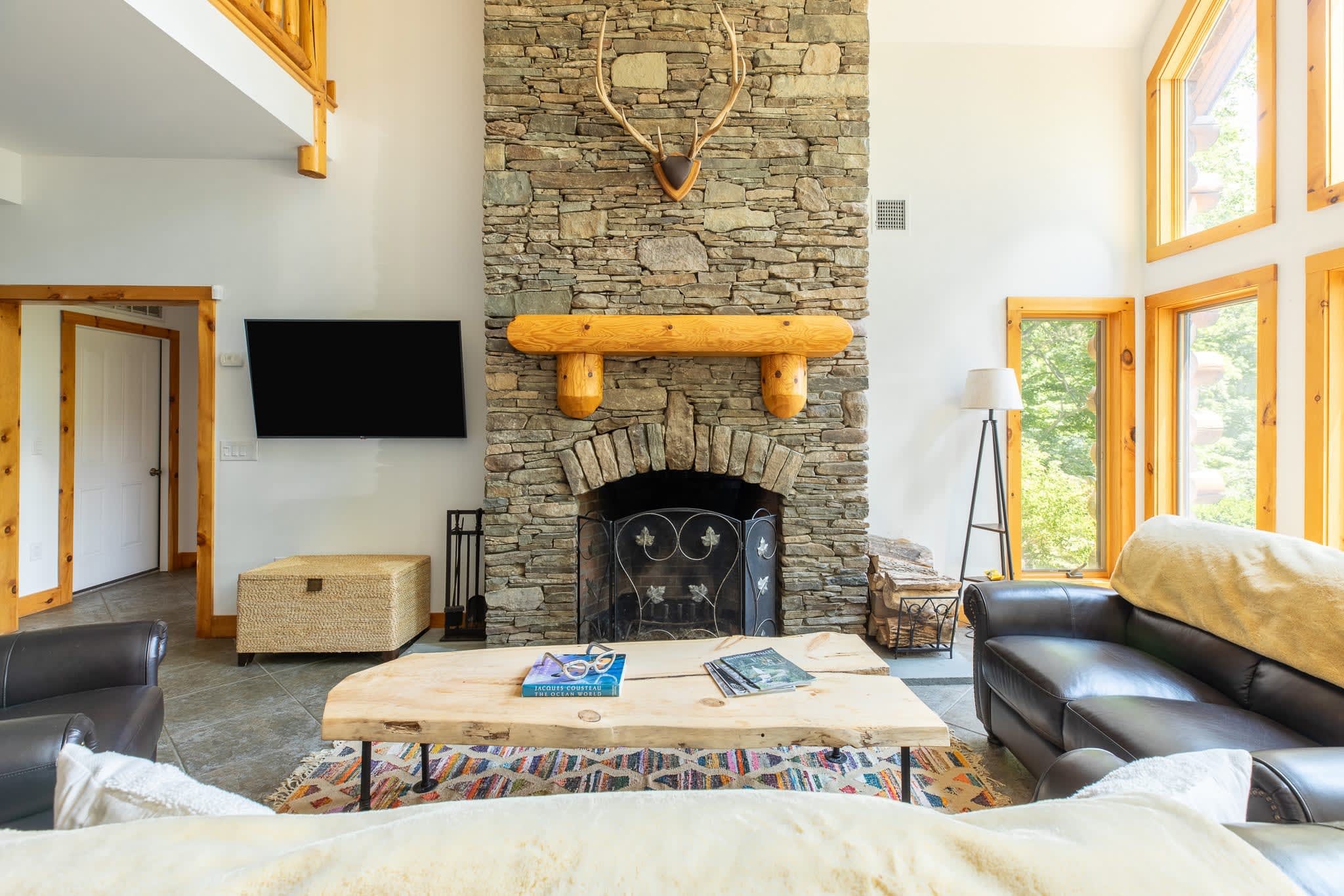 A cozy living room centers around a large stone fireplace with a rustic wooden mantel and deer antlers mounted above, complemented by a modern flat-screen TV, a live-edge wood coffee table, and dark leather seating. Large windows offer views of lush greenery.