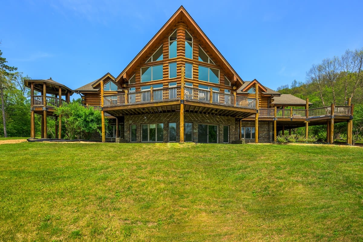 Grand log cabin lodge with a prominent A-frame design and multiple levels of windows, featuring expansive wooden decks and a stone foundation, set against a backdrop of green grass and trees.