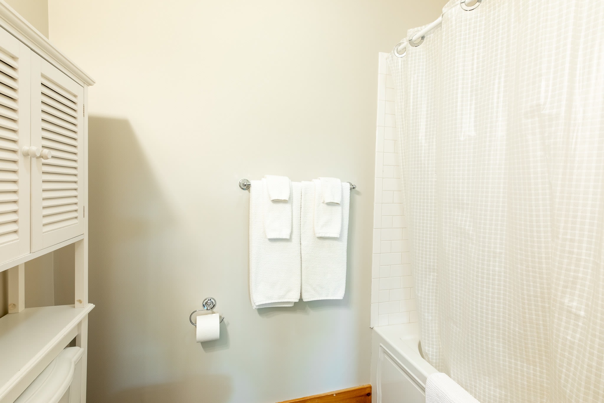 A close-up of the bathroom shows a white bathtub with a shower curtain, a white wooden cabinet over the toilet, and white towels hanging on a wall rack.