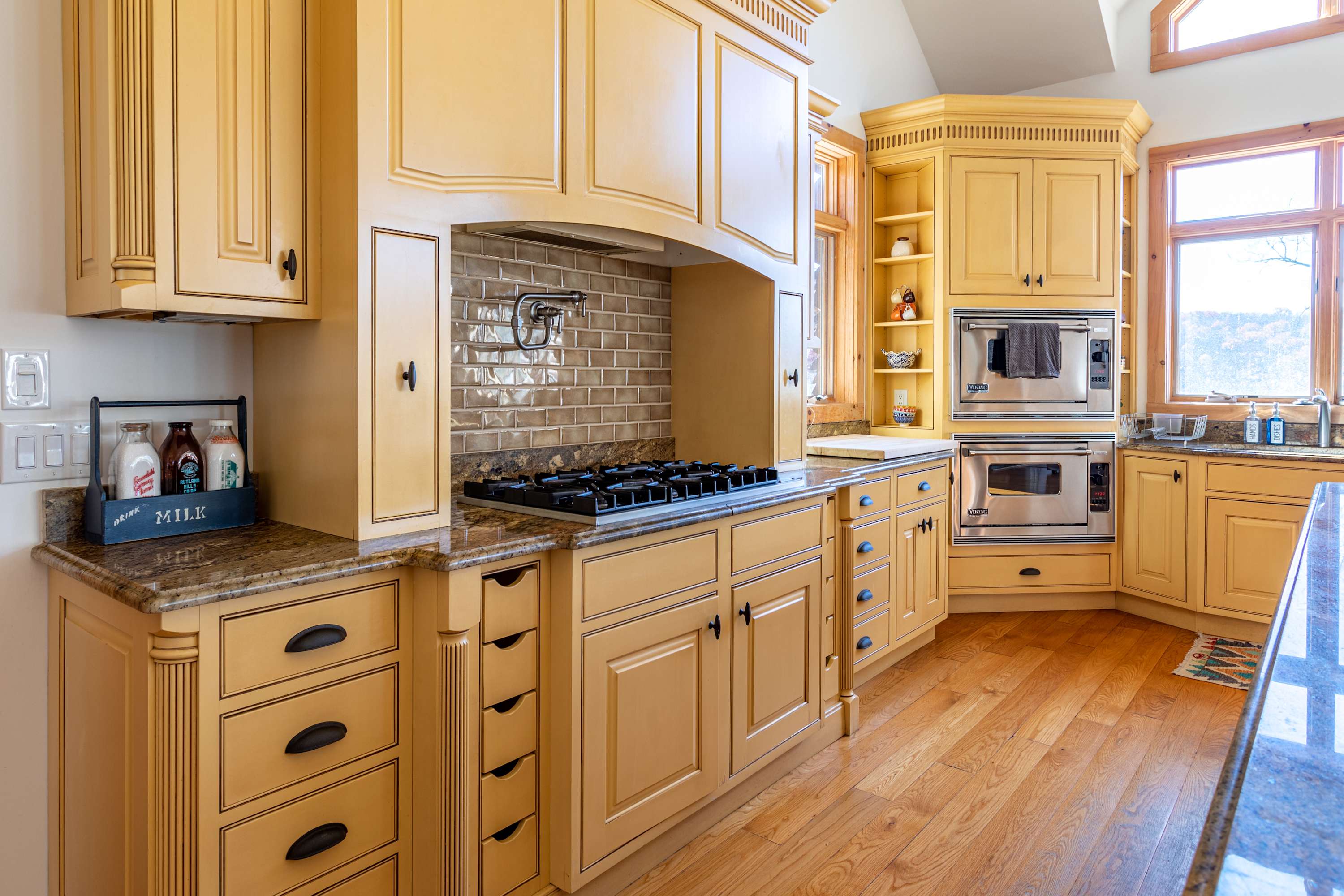 Detailed view of the kitchen's cooking area, showcasing a gas range with a subway tile backsplash and custom range hood. Light yellow cabinets and granite countertops line the walls, with built-in ovens visible to the right.