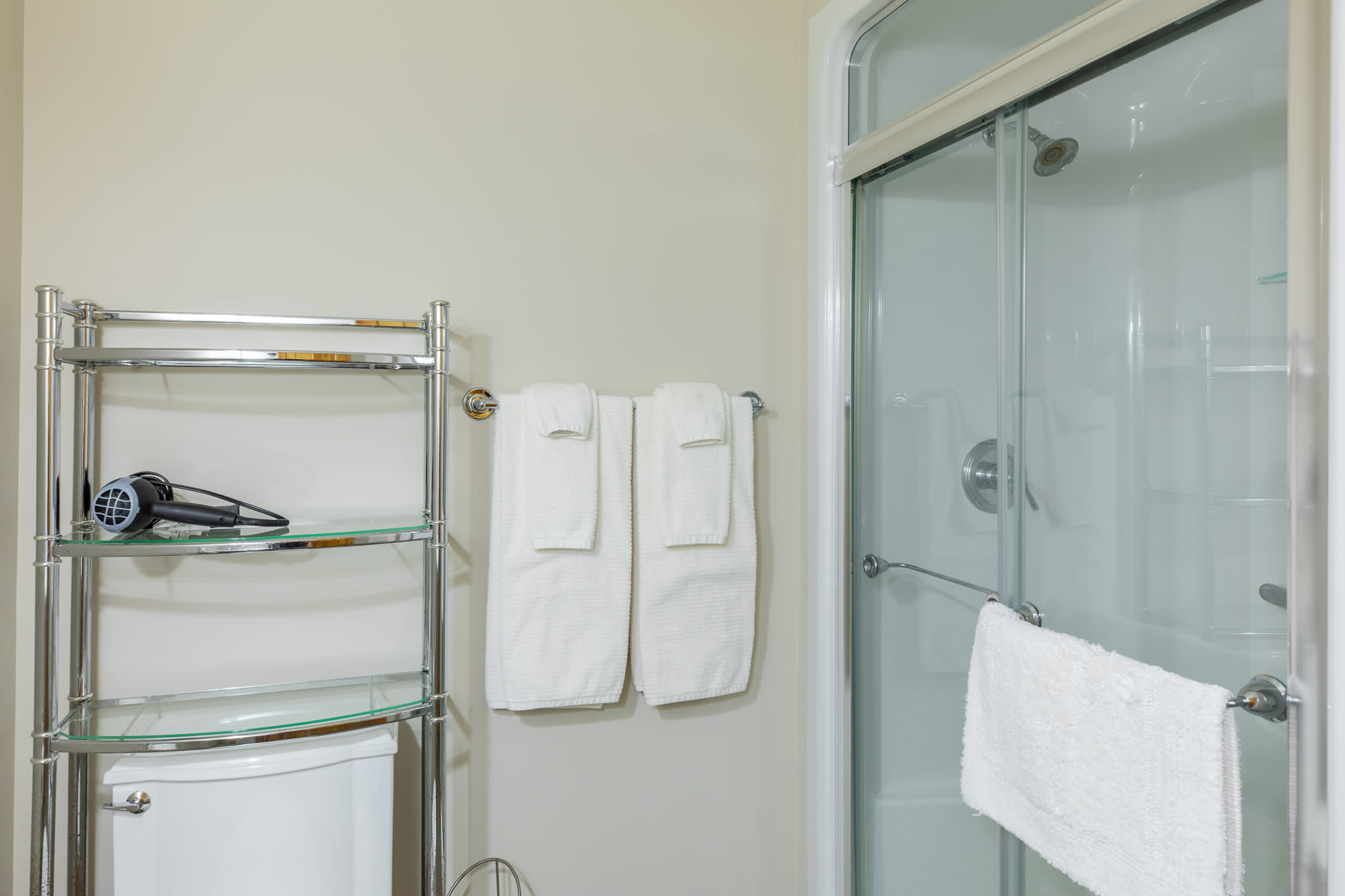 A close-up of the bathroom shows a glass-enclosed shower with a chrome fixture, a toilet, and a chrome shelving unit holding a hairdryer, with white towels neatly folded on a rack.