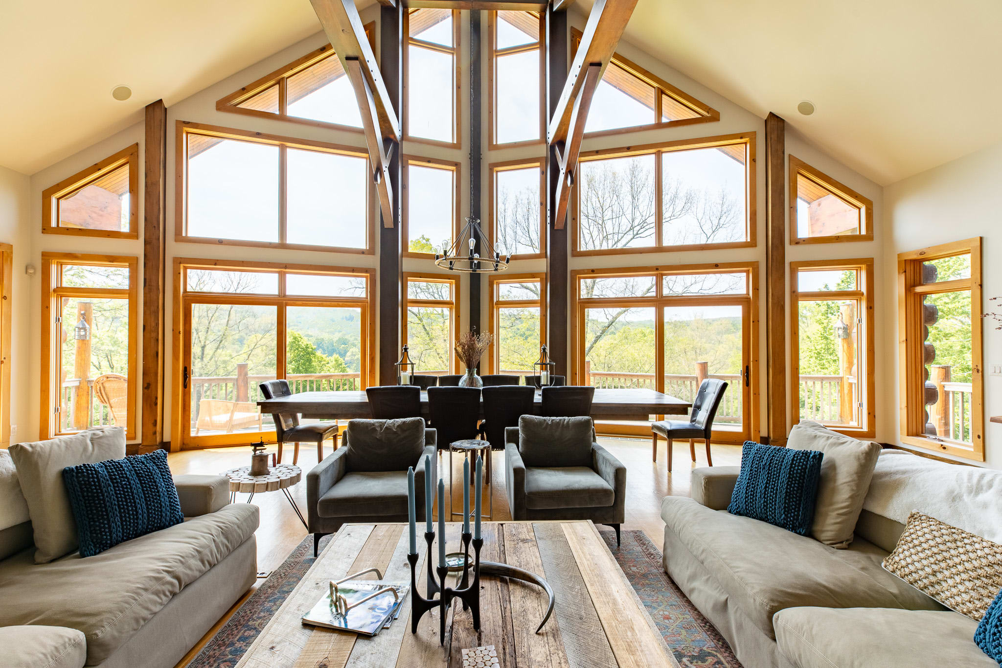 A bright and airy living room features two light-colored sofas and two grey armchairs facing a rustic wooden coffee table, with a view towards the expansive dining area and floor-to-ceiling windows offering scenic outdoor views. Exposed wooden beams frame the vaulted ceiling.