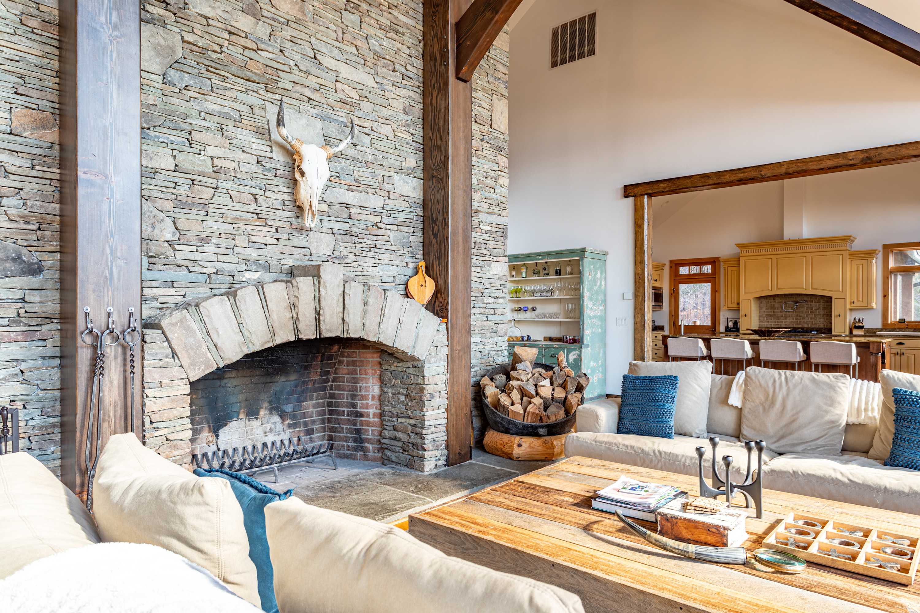 A cozy living area features a large stone fireplace with a prominent animal skull decoration above the mantel, flanked by light-colored sofas with blue accent pillows. A rustic wooden coffee table sits on a patterned rug, with a glimpse of the kitchen beyond.