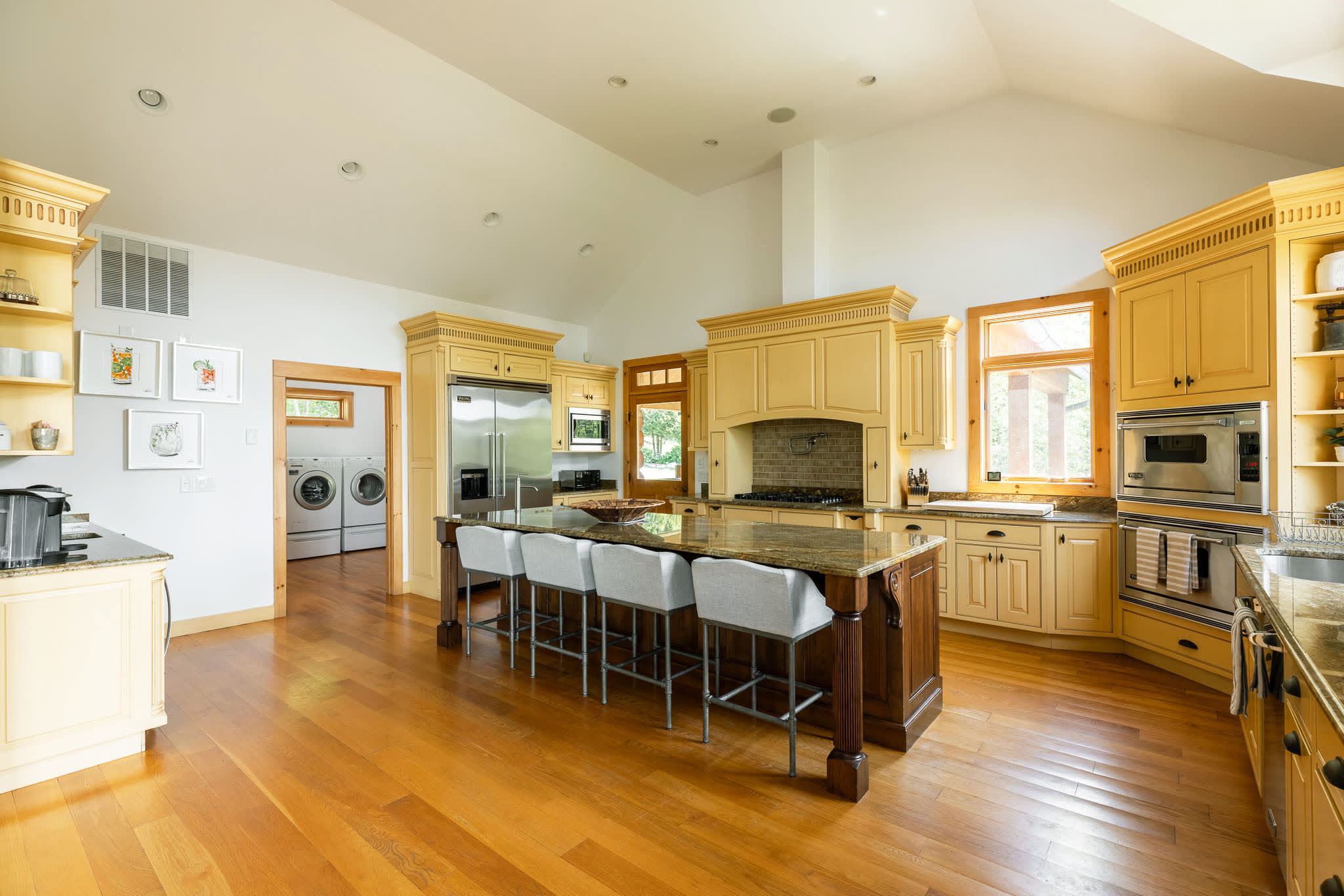 Expansive kitchen with vaulted ceilings, a large central island, and light yellow custom cabinetry. Stainless steel appliances are integrated, and a doorway leads to a separate laundry room with a washer and dryer.