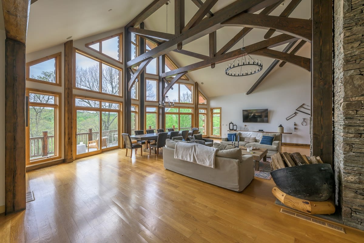 Expansive open-concept living and dining area featuring vaulted ceilings with exposed timber beams, large windows offering lush green views, and a stone fireplace. Light-colored sofas define the living space, while a long dining table is visible in the background.