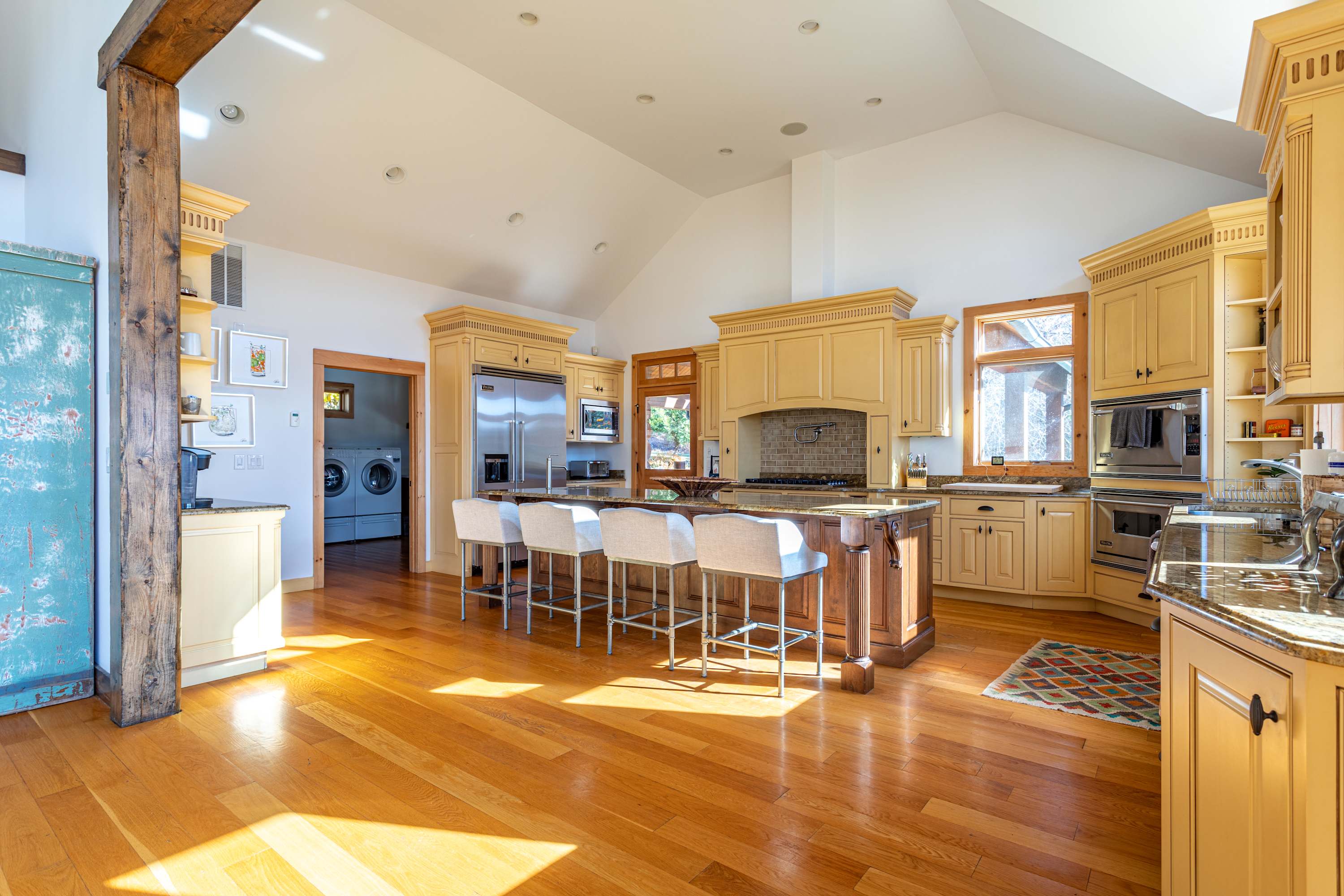 Bright and airy kitchen with vaulted ceilings, a large central island with seating, and extensive light yellow cabinetry. A doorway in the background reveals a glimpse of a laundry area with a washer and dryer.
