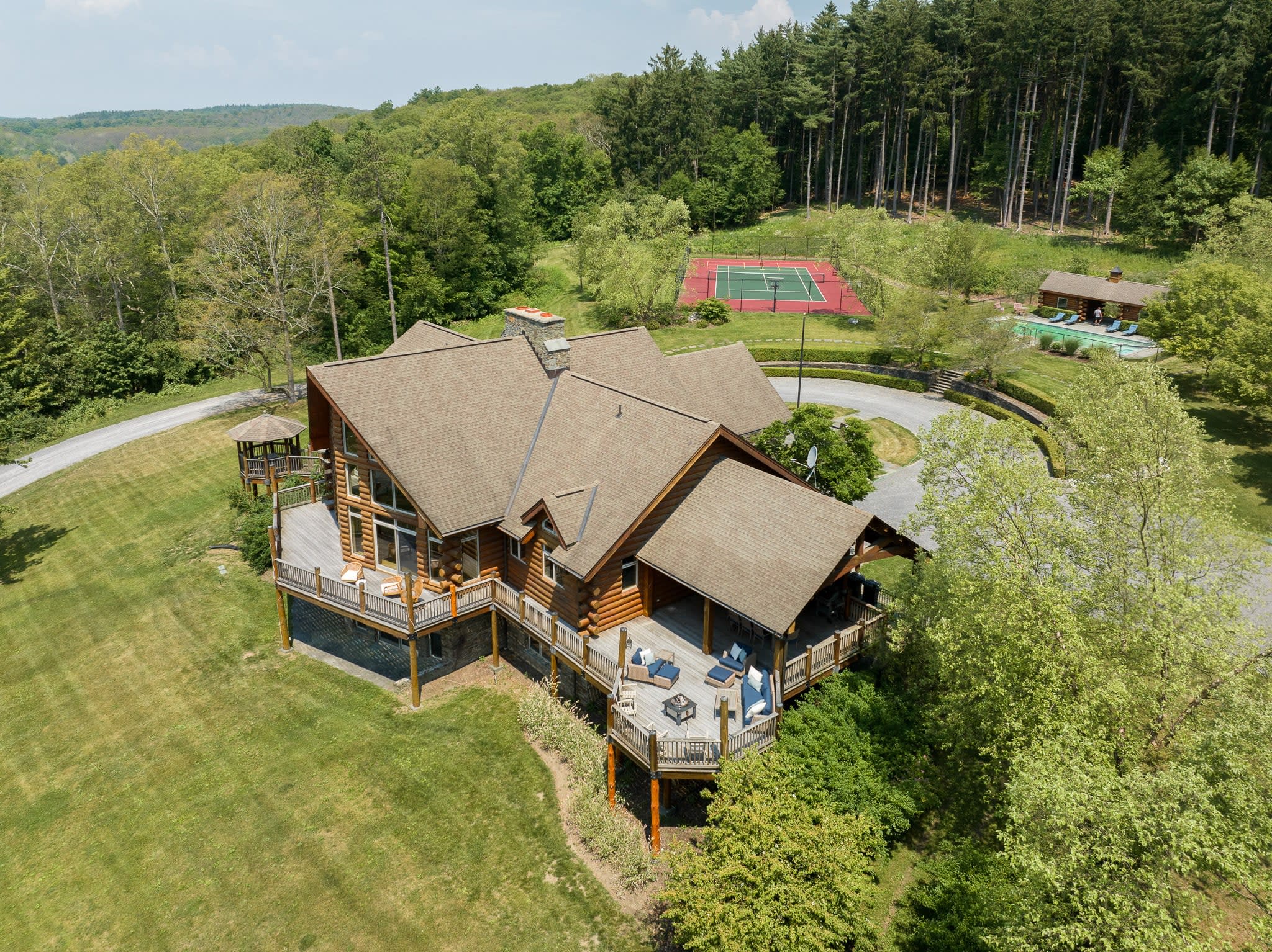 Aerial view of the large log cabin lodge with multiple gables and extensive wooden decks, surrounded by green lawns and trees, with a tennis court and pool area visible in the distance.
